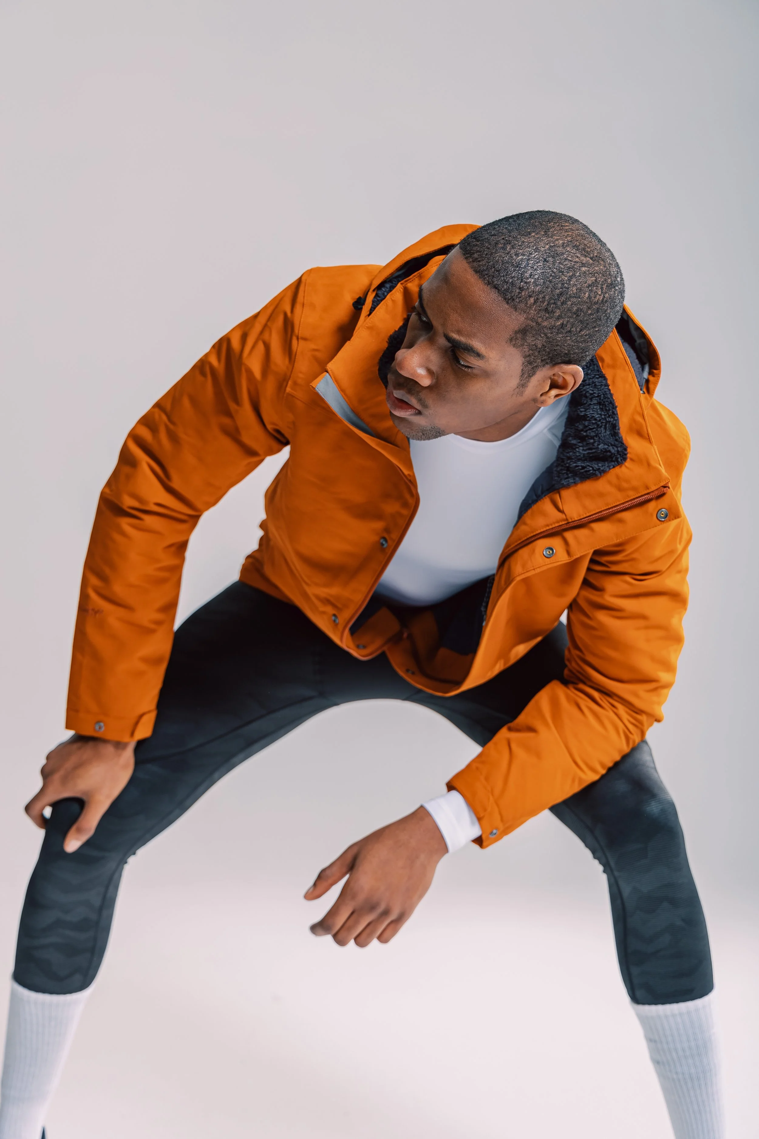 A young man in an orange jacket, white shirt, black patterned athletic pants, and white socks is in a squatting position, leaning forward with his hands resting on his knees, against a plain light background.
