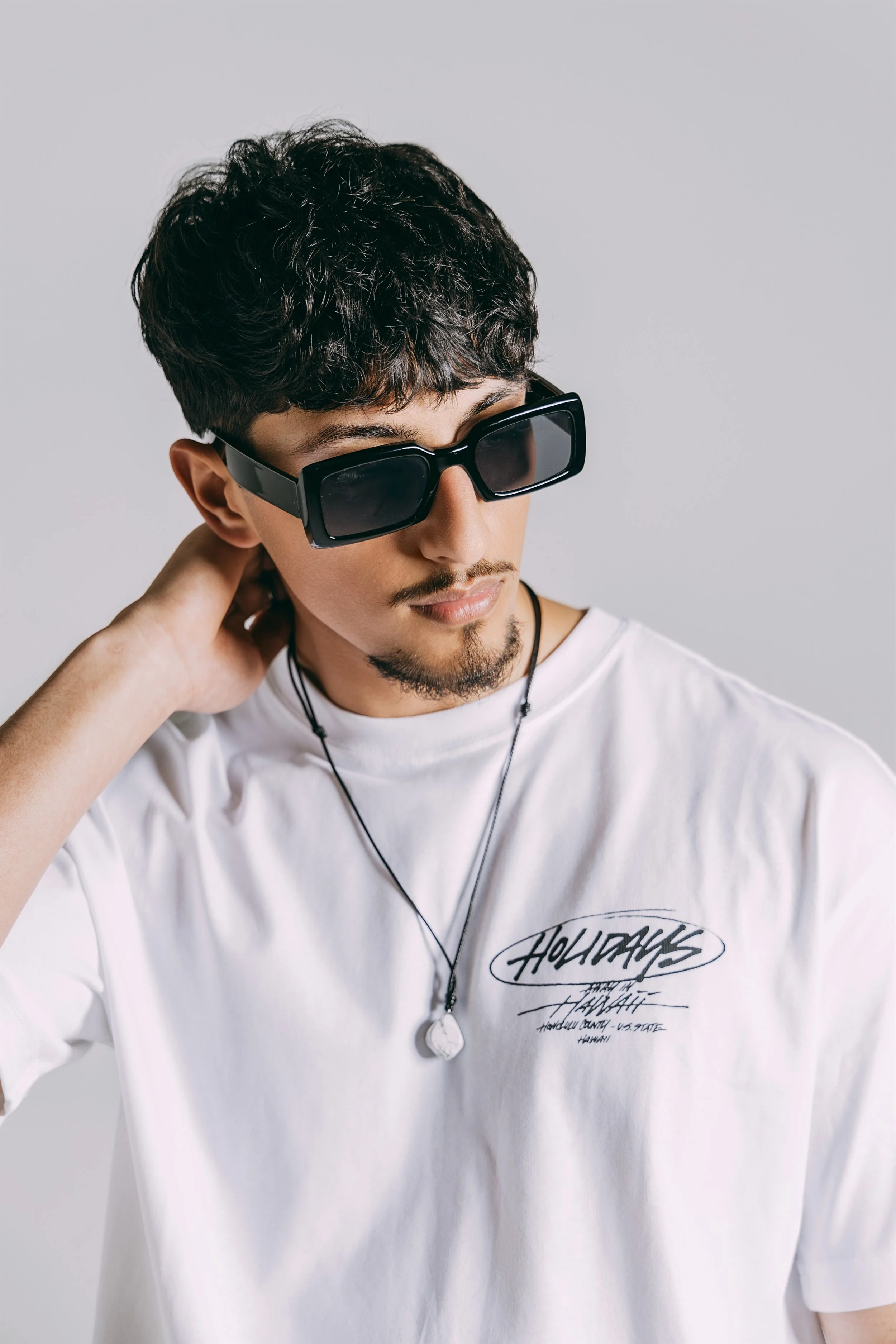 A young man with dark curly hair, a goatee, and mustache wearing oversized sunglasses, a white T-shirt with a graphic, and a necklace, posing with one hand behind his neck against a plain background.