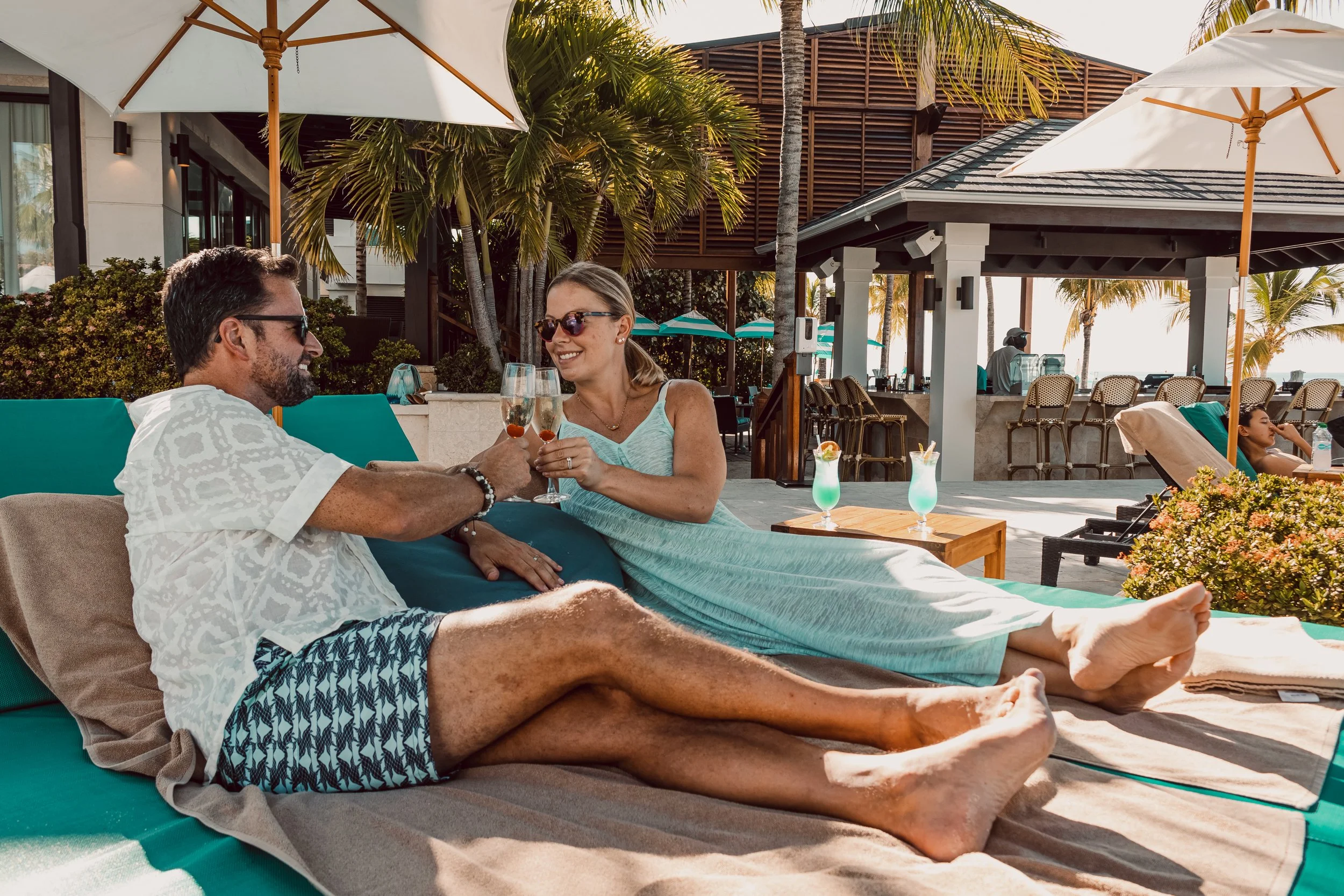 A man and a woman sitting by a pool, smiling and clinking glasses, with white umbrellas, palm trees, and a building in the background.