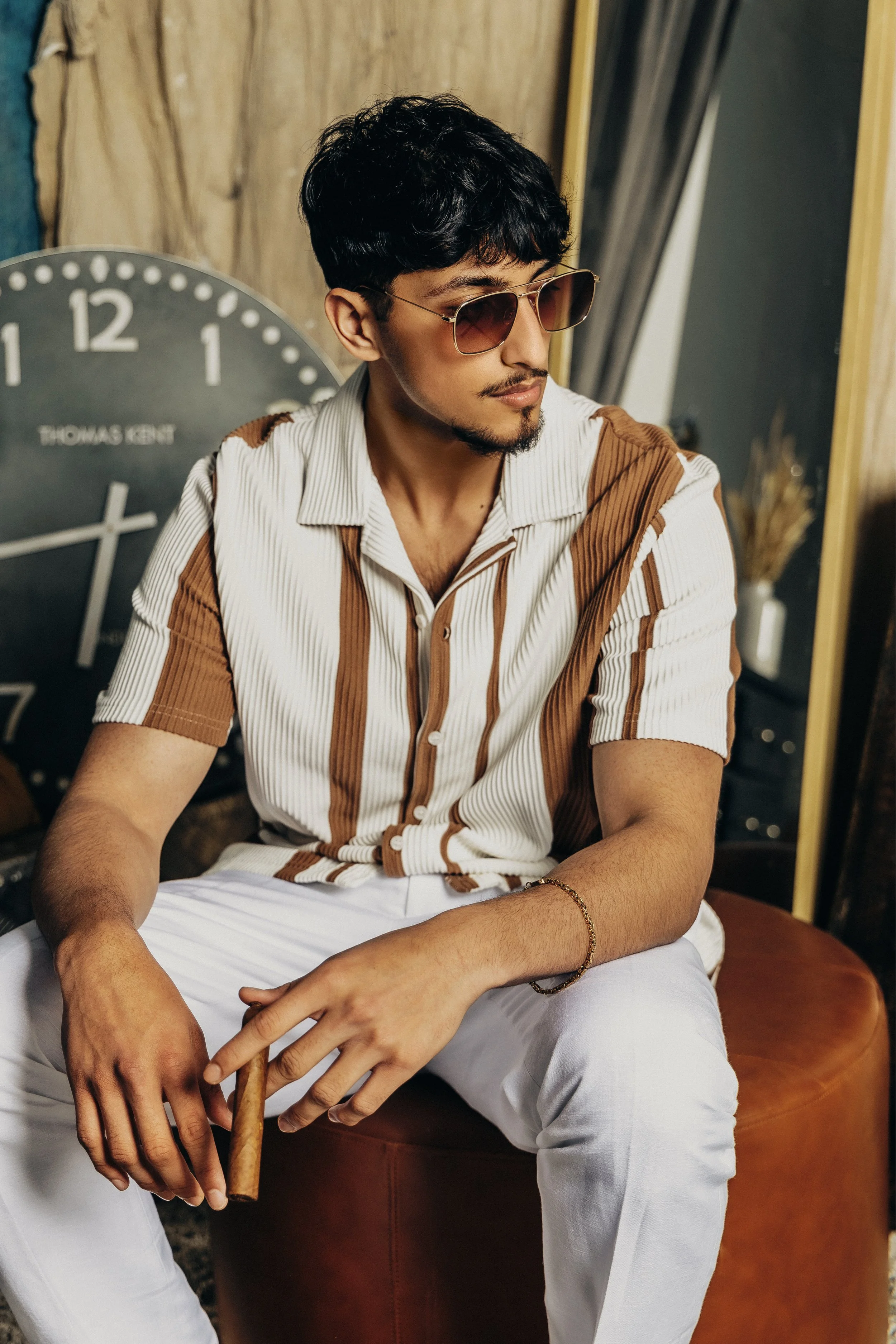 A young man with dark hair, wearing sunglasses, a striped short-sleeve shirt, and white pants, sitting on a brown leather ottoman in a room with a large clock and decorative items in the background.