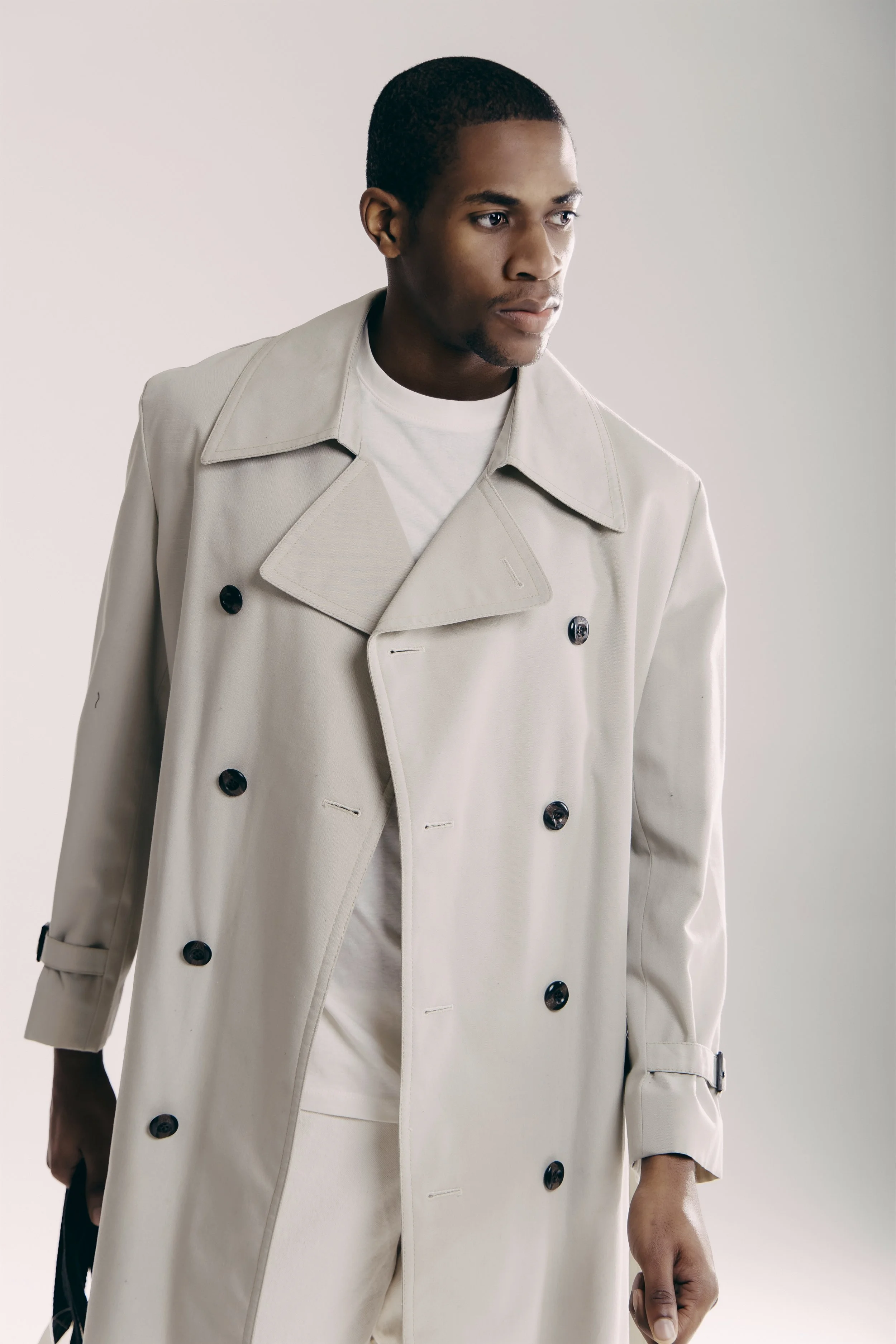 A young man with dark skin and short hair wears a beige trench coat over a white shirt, standing against a plain light background.