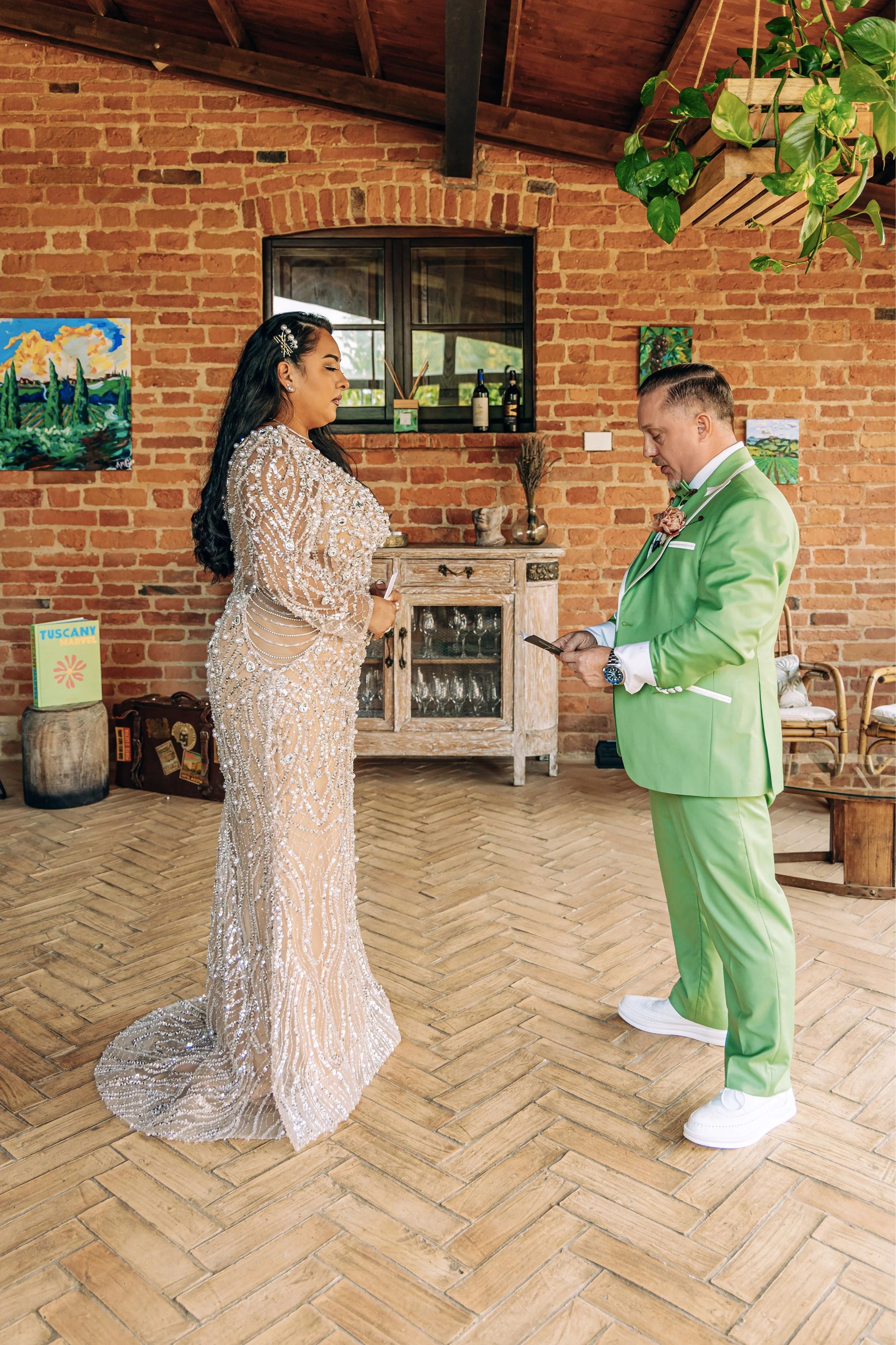 A woman in an elaborate, sparkling wedding dress facing a man in a bright green suit, both standing in a rustic room with brick walls and wooden floors. The woman has long dark hair and is wearing a hair accessory, while the man is looking at his pho