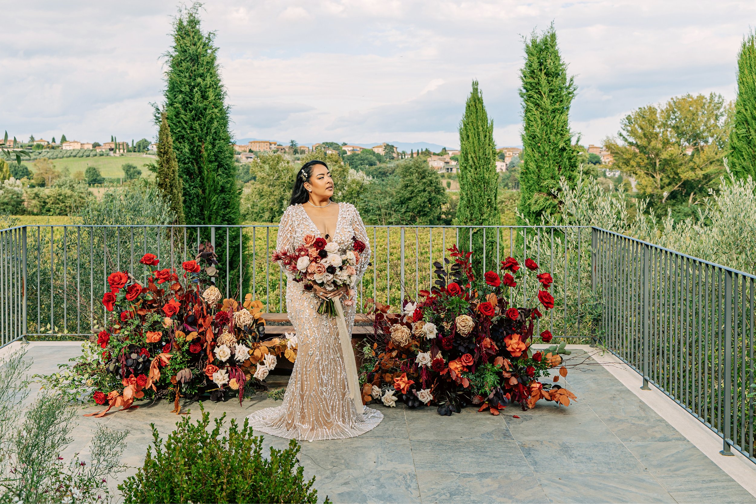 A woman wearing an ornate, sparkling gown stands on a balcony adorned with vibrant red and beige flowers, with a scenic landscape of trees and hills in the background.