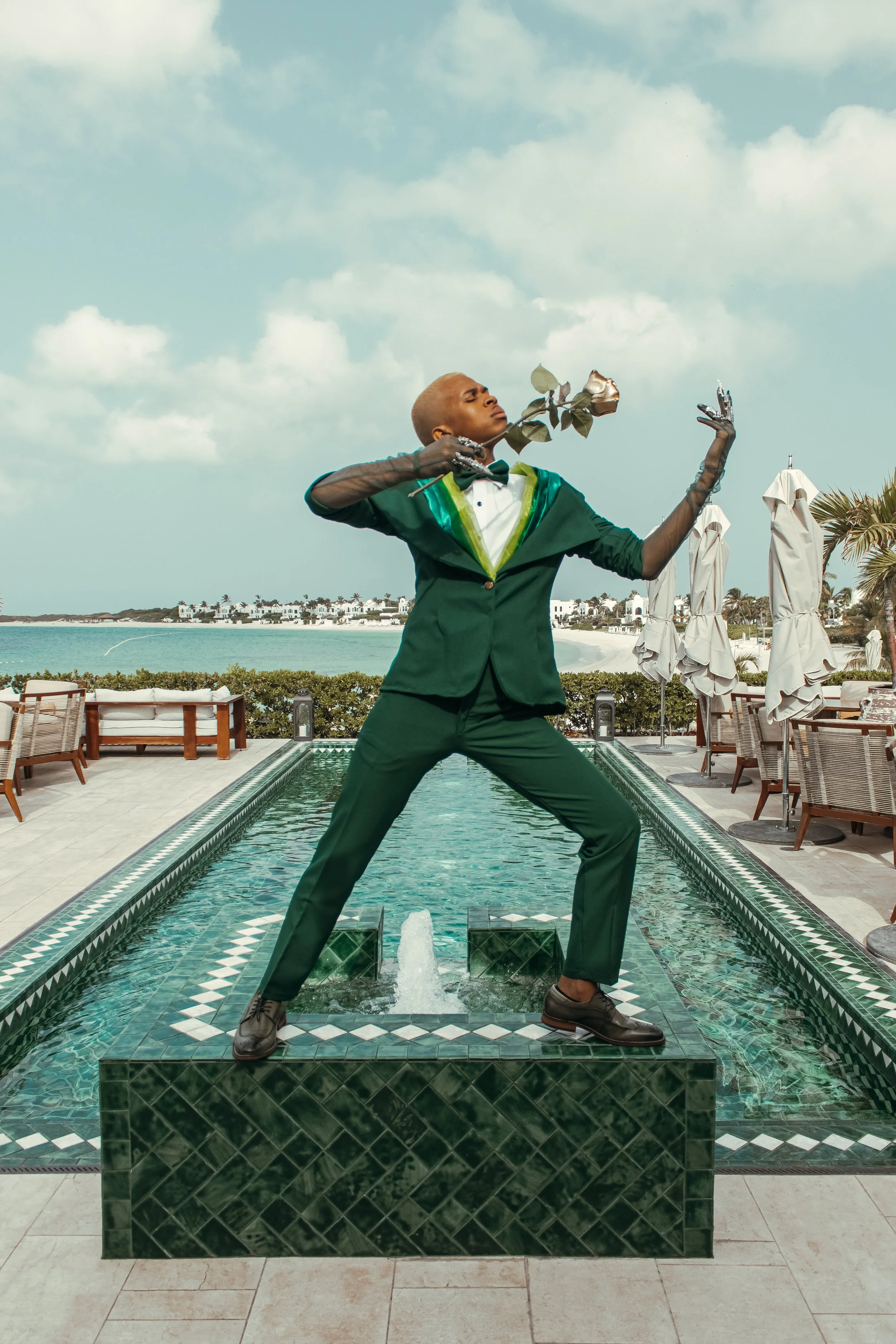 Person in a green suit with a tuxedo shirt standing on a fountain at an outdoor poolside setting, holding a silver rose and striking a pose.