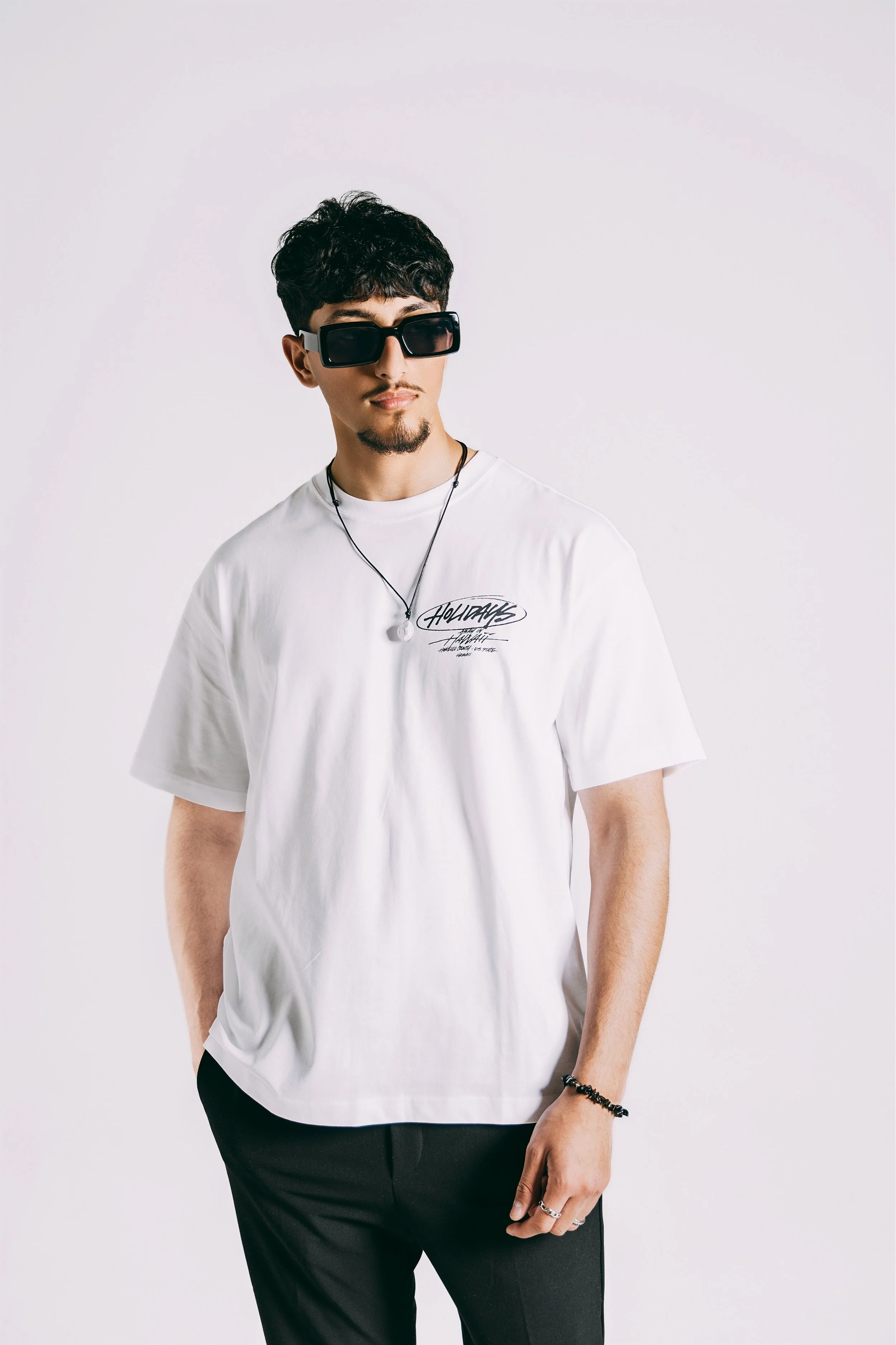 A young man with dark hair, sunglasses, and a white graphic T-shirt posing against a plain white background.