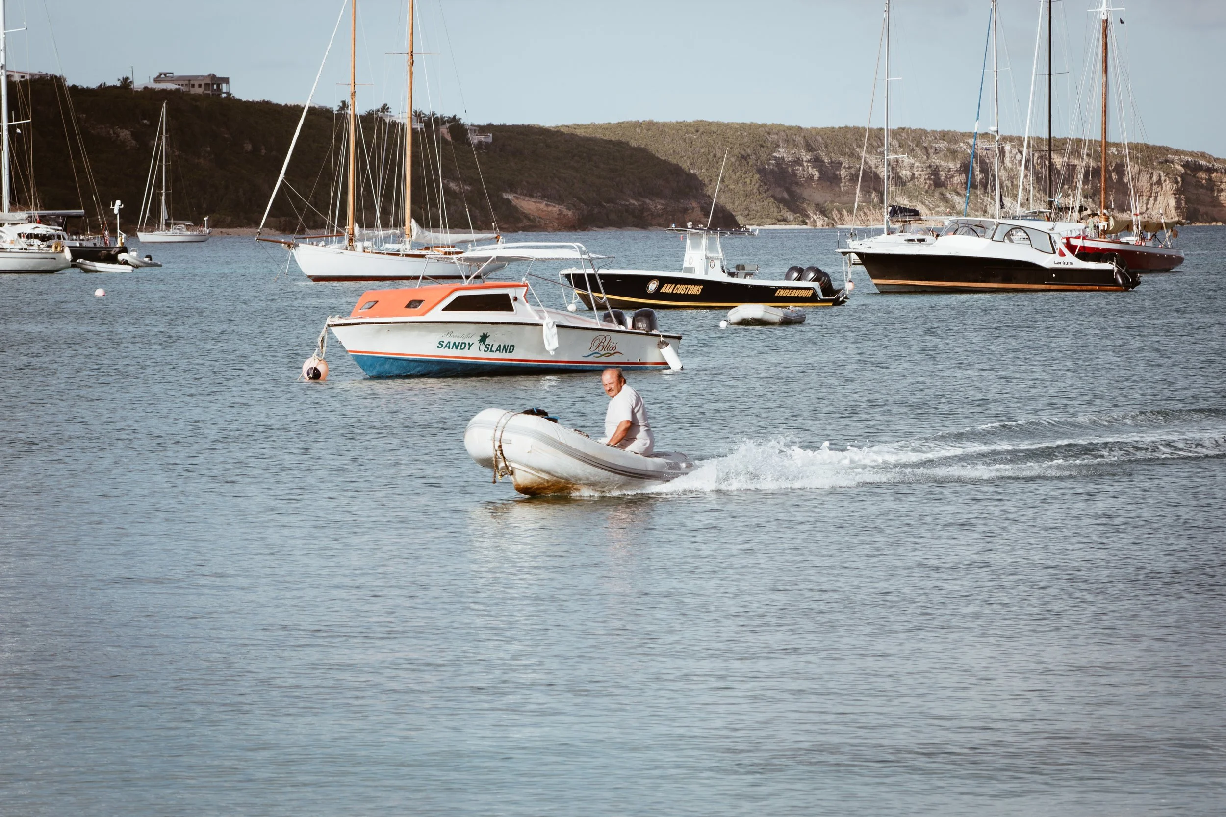 A man in a white shirt is riding a small motorized inflatable boat on the water near several anchored sailboats with a hilly shoreline in the background.