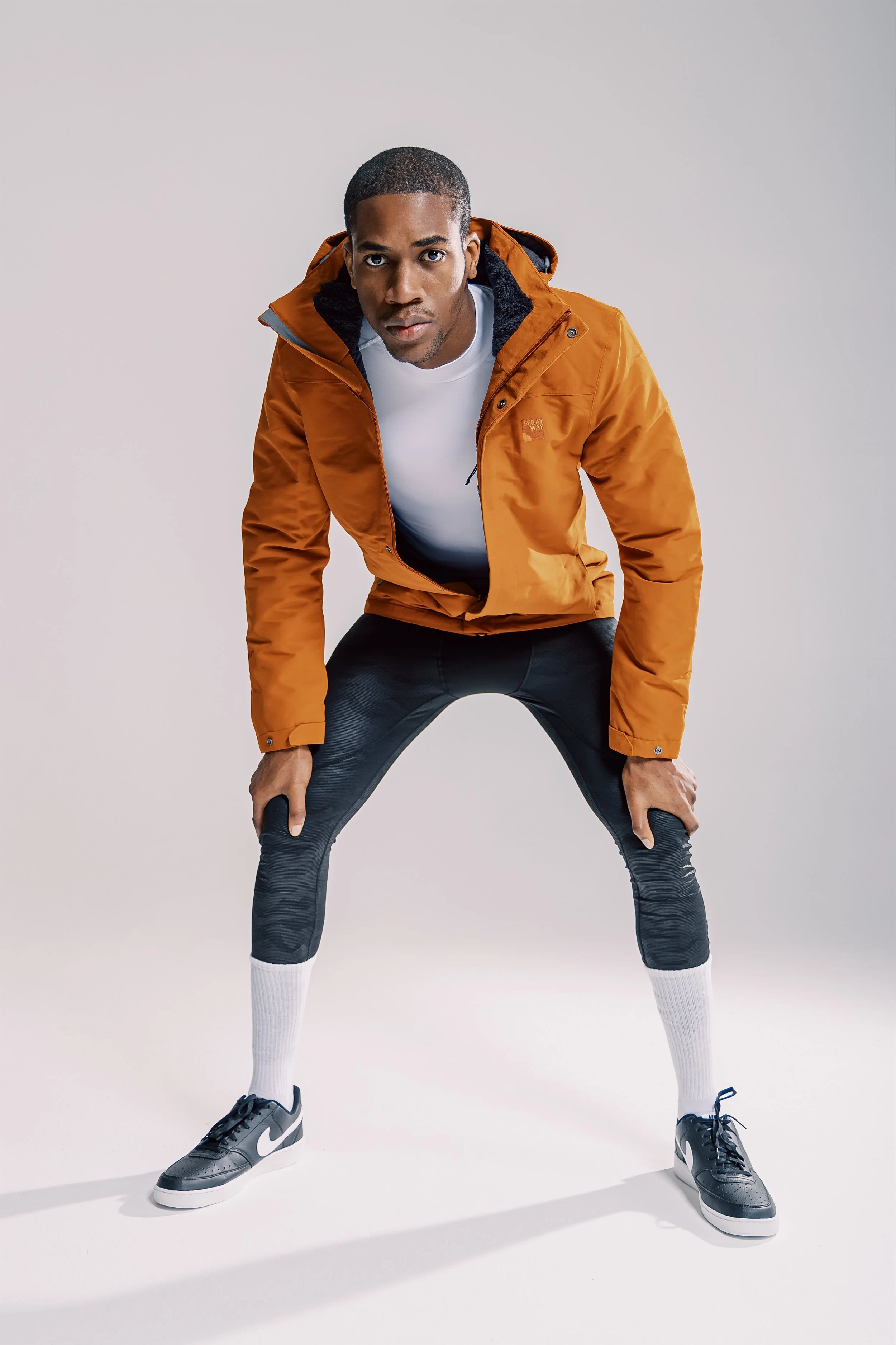 Man in orange jacket, white shirt, black pants, and sneakers in a wide stance on a white background.