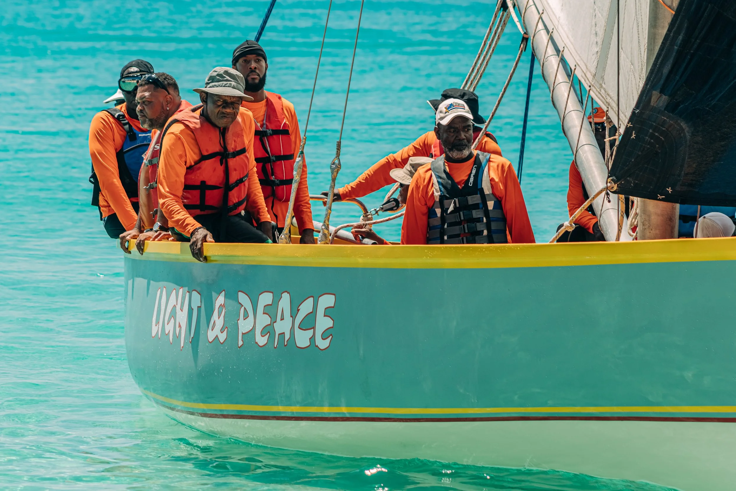 A group of diverse people wearing orange life jackets on a yellow sailboat on turquoise water, with the words 'Light & Peace' written on the boat's side.
