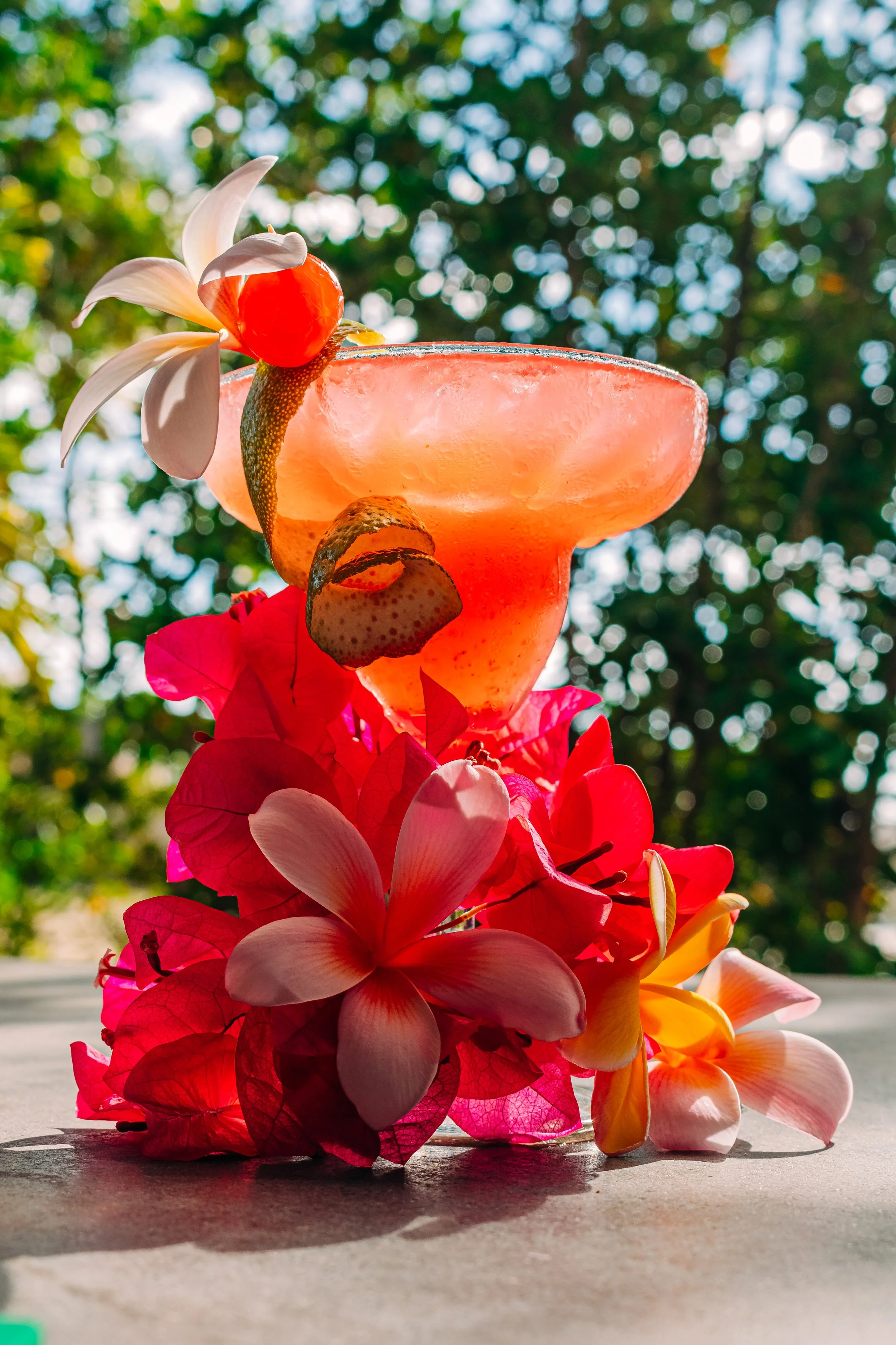 A colorful tropical cocktail garnished with a cherry, white flower, and citrus peel, decorated with pink and red flowers, on a surface outdoors with a blurred green leafy background.