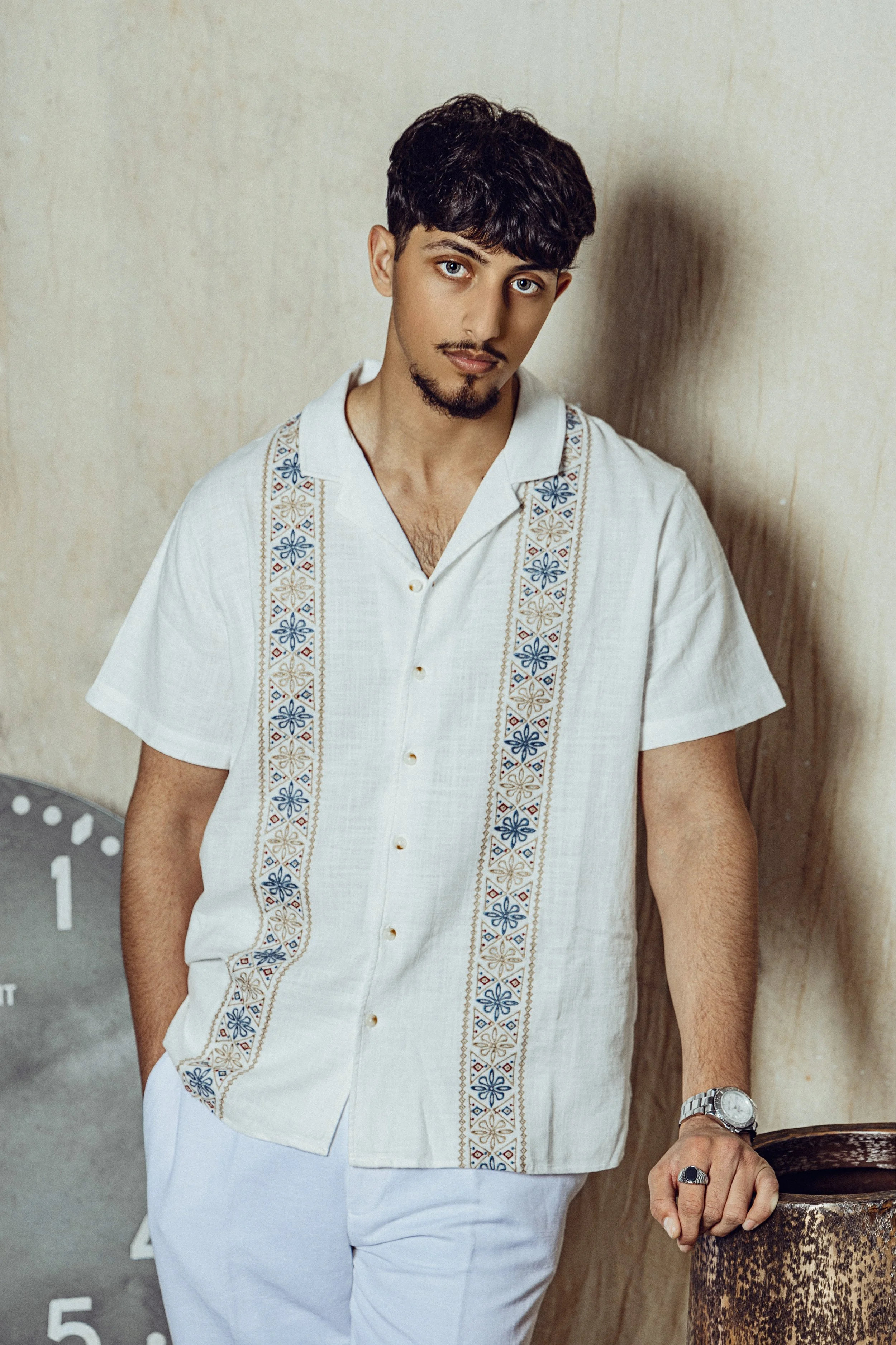A young man with dark, curly hair, blue eyes, a mustache, and a goatee, wearing a white short-sleeve shirt with embroidered patterns on the front. He has a silver watch on his left wrist and a black ring on his right hand. He is leaning against a tex