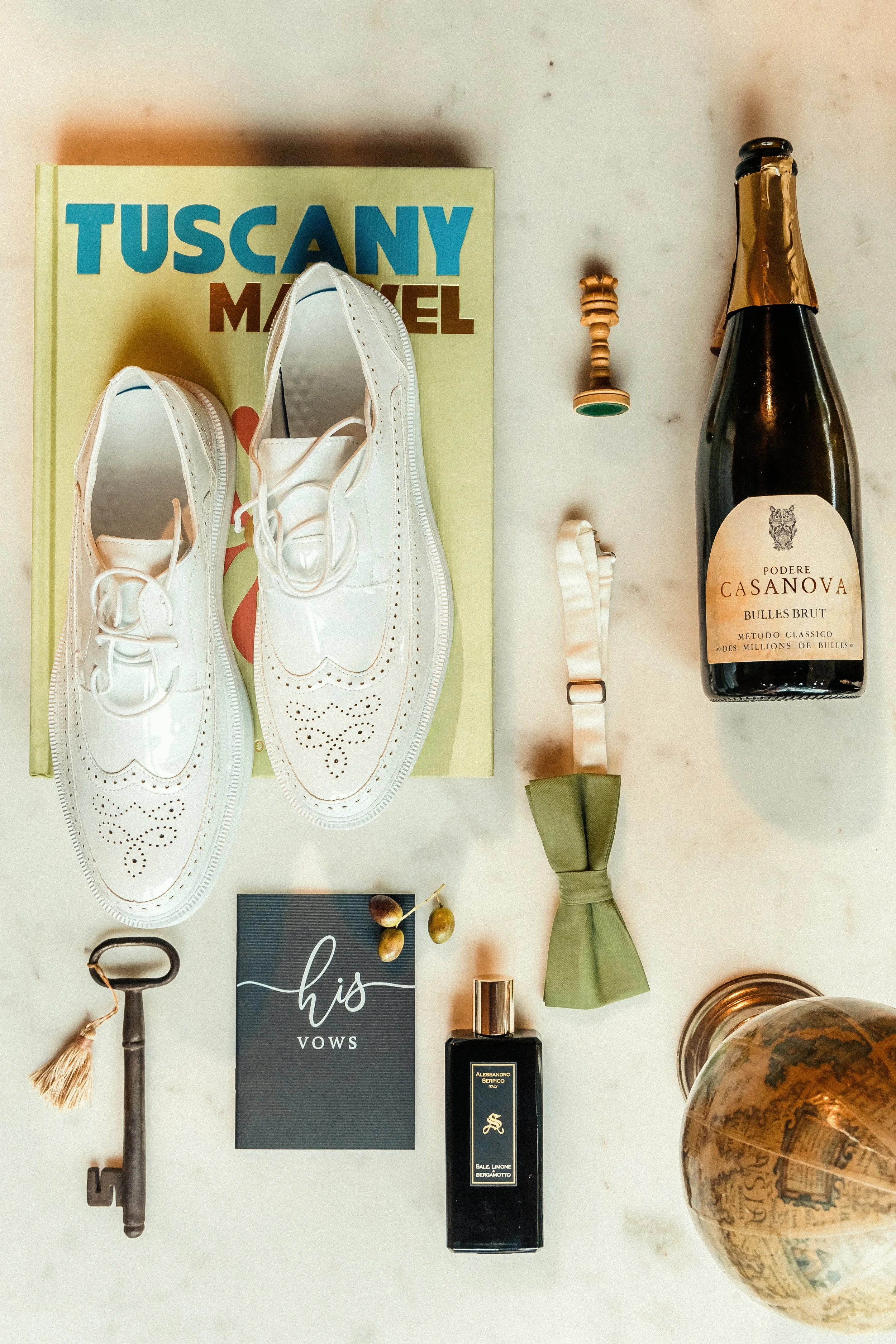 Flat lay of a book titled 'Tuscany', white shoes, a wine bottle labeled 'Podere Casanova', a green bow tie, a black bottle of perfume, a vintage key, a card that says 'his vows', a small chess piece, and a glass bowl on a marble surface.