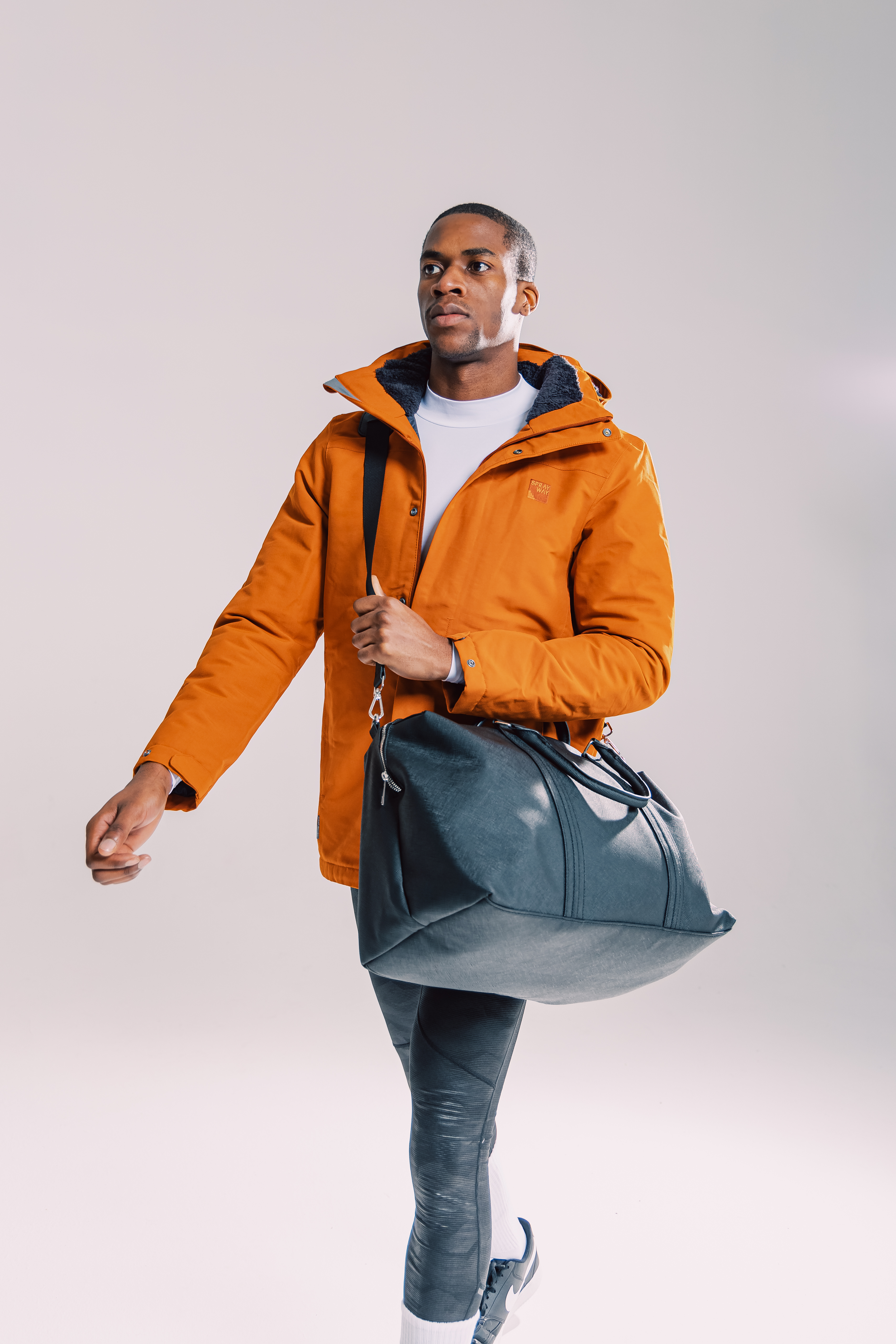 A man dressed in an orange jacket, light gray sports pants, and sneakers, carrying a black gym bag over his shoulder, walking forward.