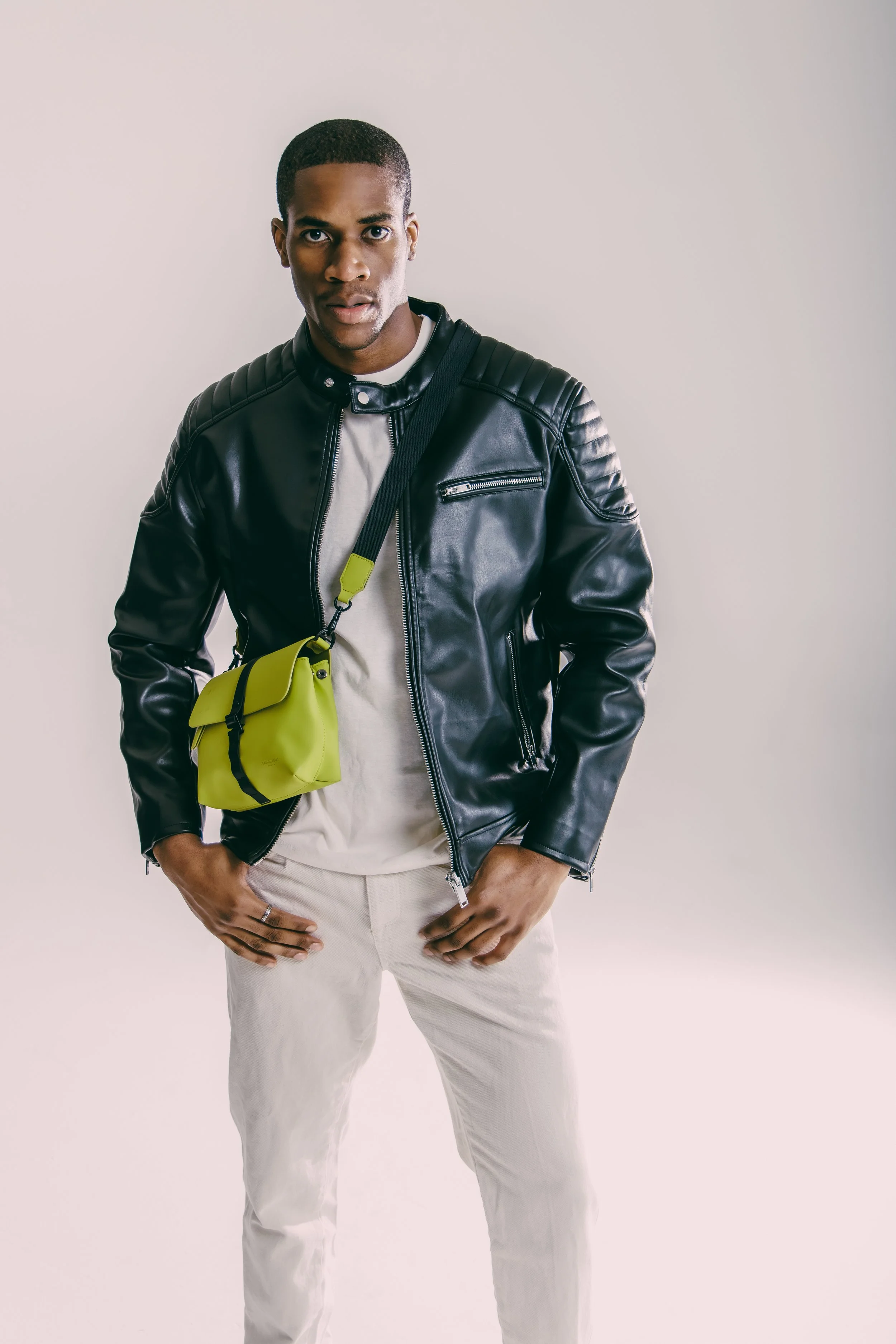 Young man with a serious expression, wearing a black leather jacket, beige pants, and a small yellow crossbody bag, standing against a plain white background.