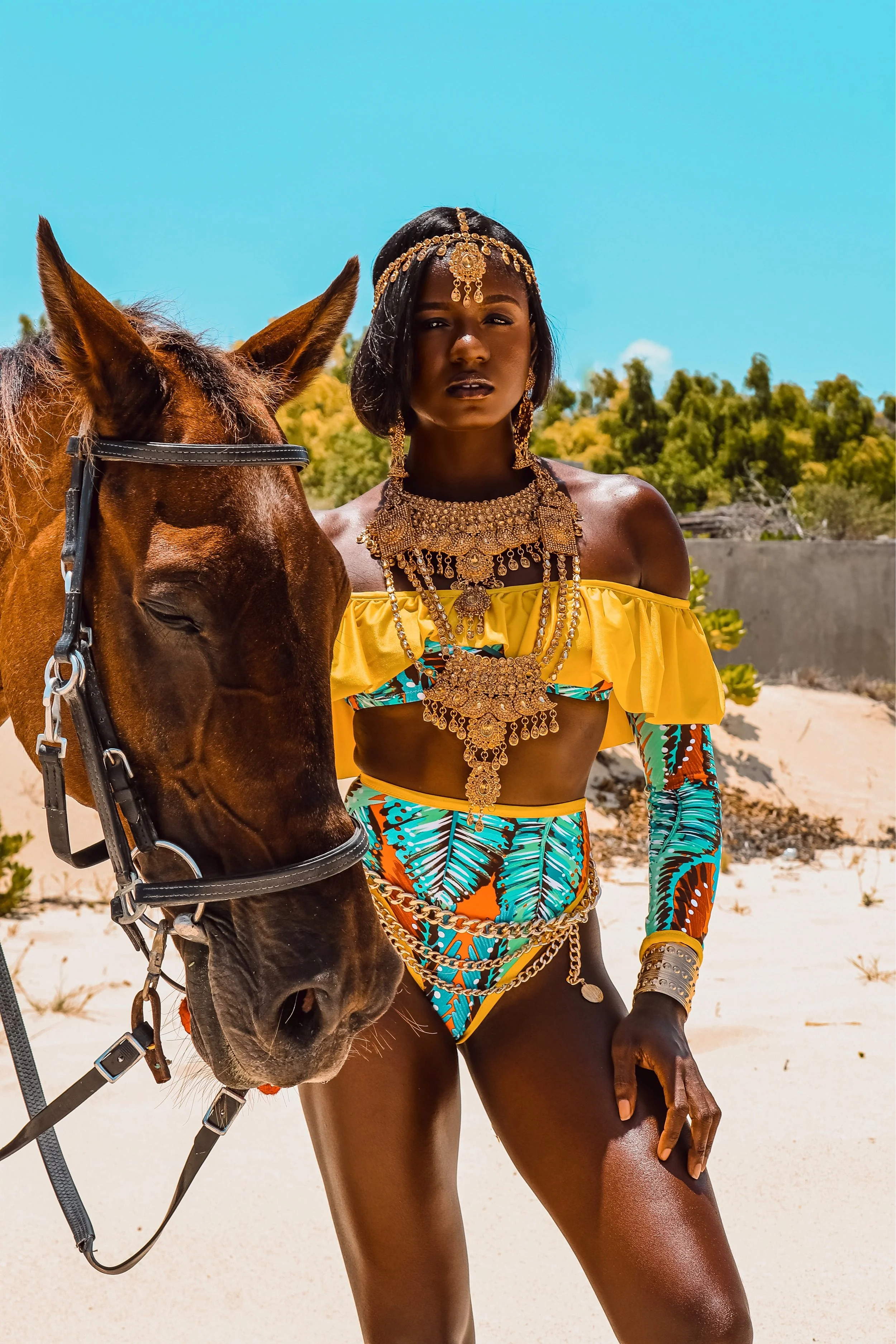 A woman dressed in colorful, revealing clothing and elaborate jewelry standing next to a brown horse on a sandy, desert-like landscape with trees and a blue sky in the background.