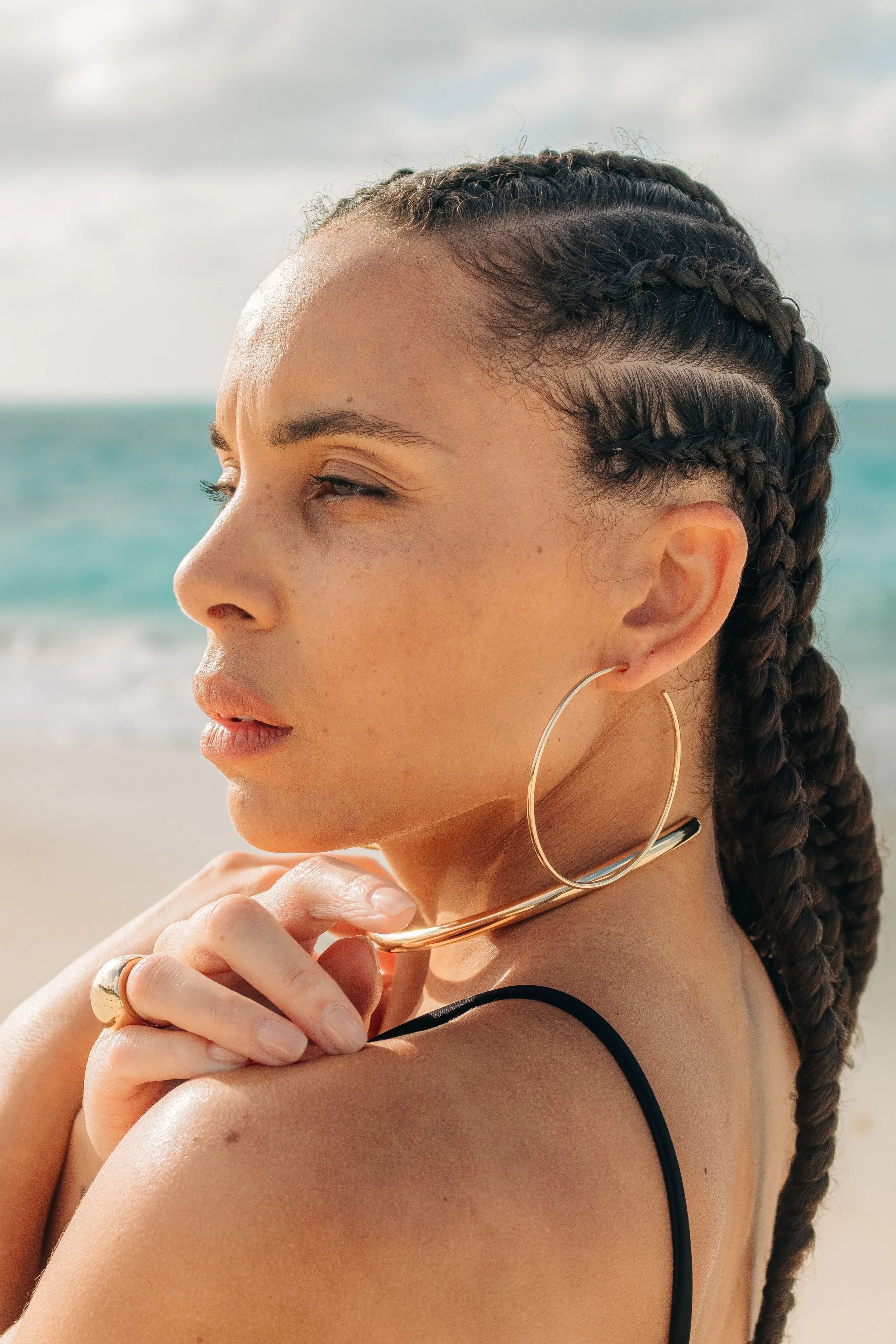 Close-up of a young woman with braided hair and hoop earrings on a beach with the ocean and cloudy sky in the background.