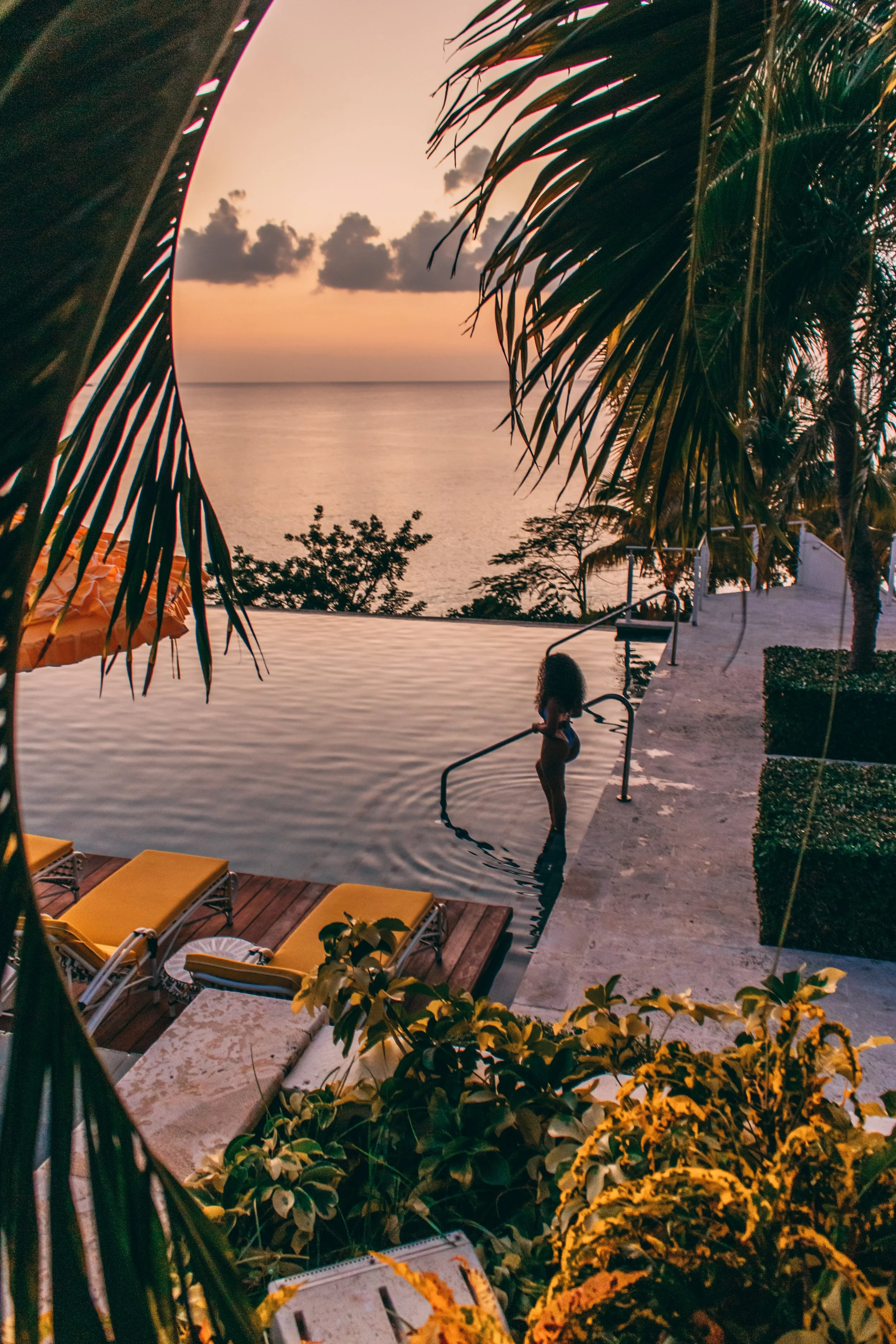 A woman in a swimsuit standing in a swimming pool at sunset, with tropical trees and clouds in the background, and poolside lounge chairs with yellow cushions in the foreground.