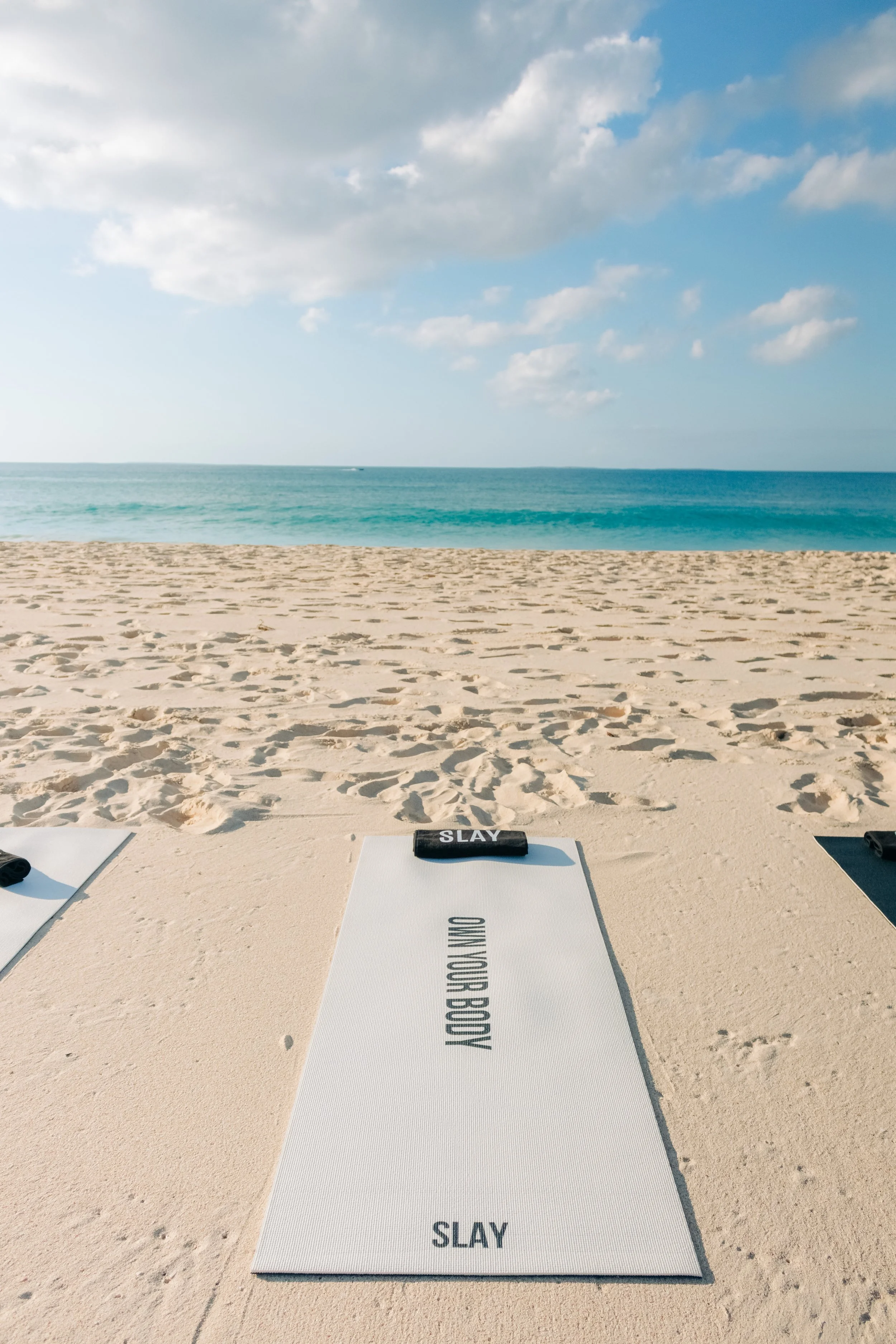 Yoga mat on the sandy beach with the ocean and cloudy sky in the background. The mat says 'OMO YOGA BOJI' and has a black towel with 'SLAY' written on it placed on top.