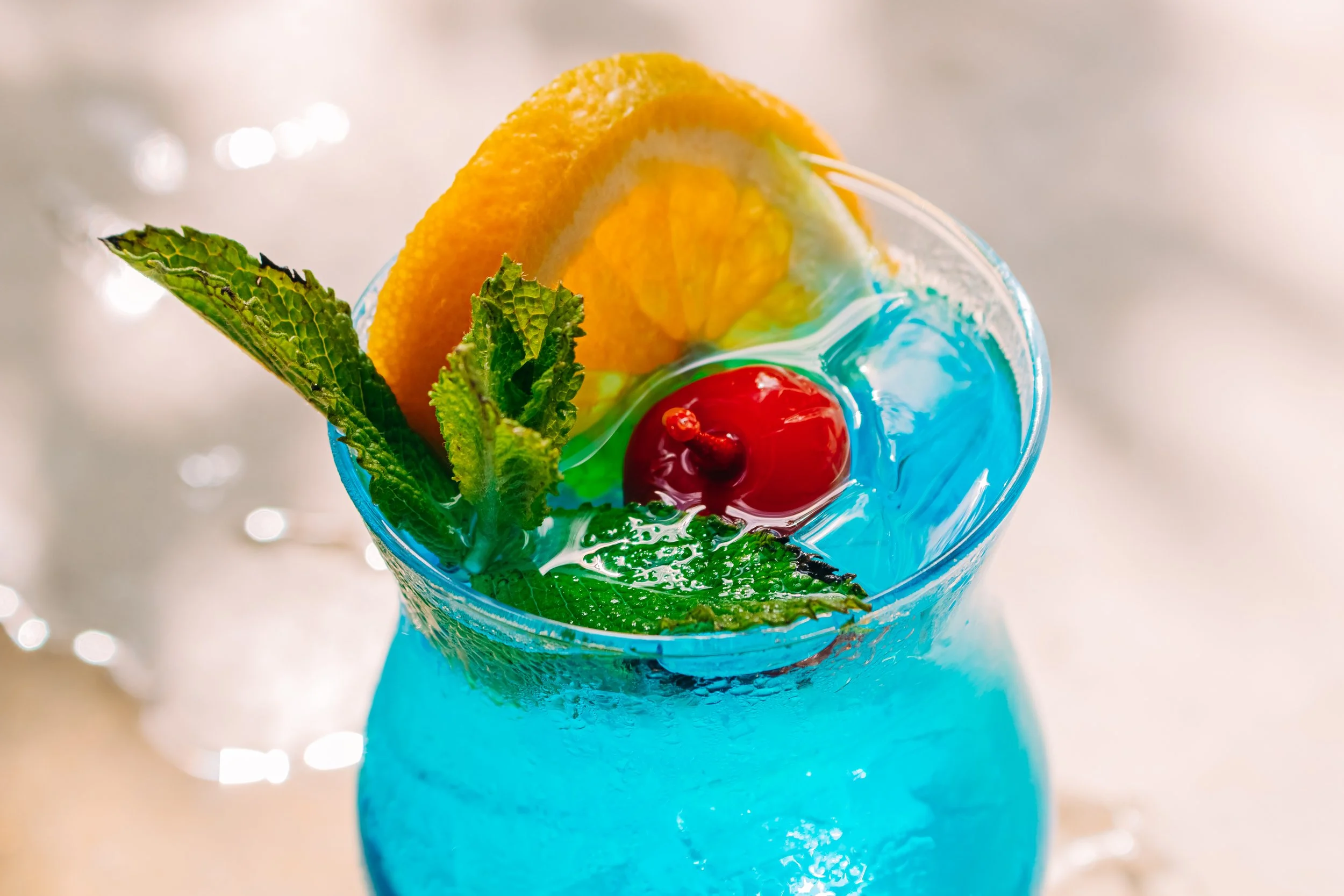 A vibrant, colorful tropical cocktail topped with orange slices, cherries, fresh mint leaves, and decorative blue and green swirled sugar or syrup, served in a tall, textured glass.