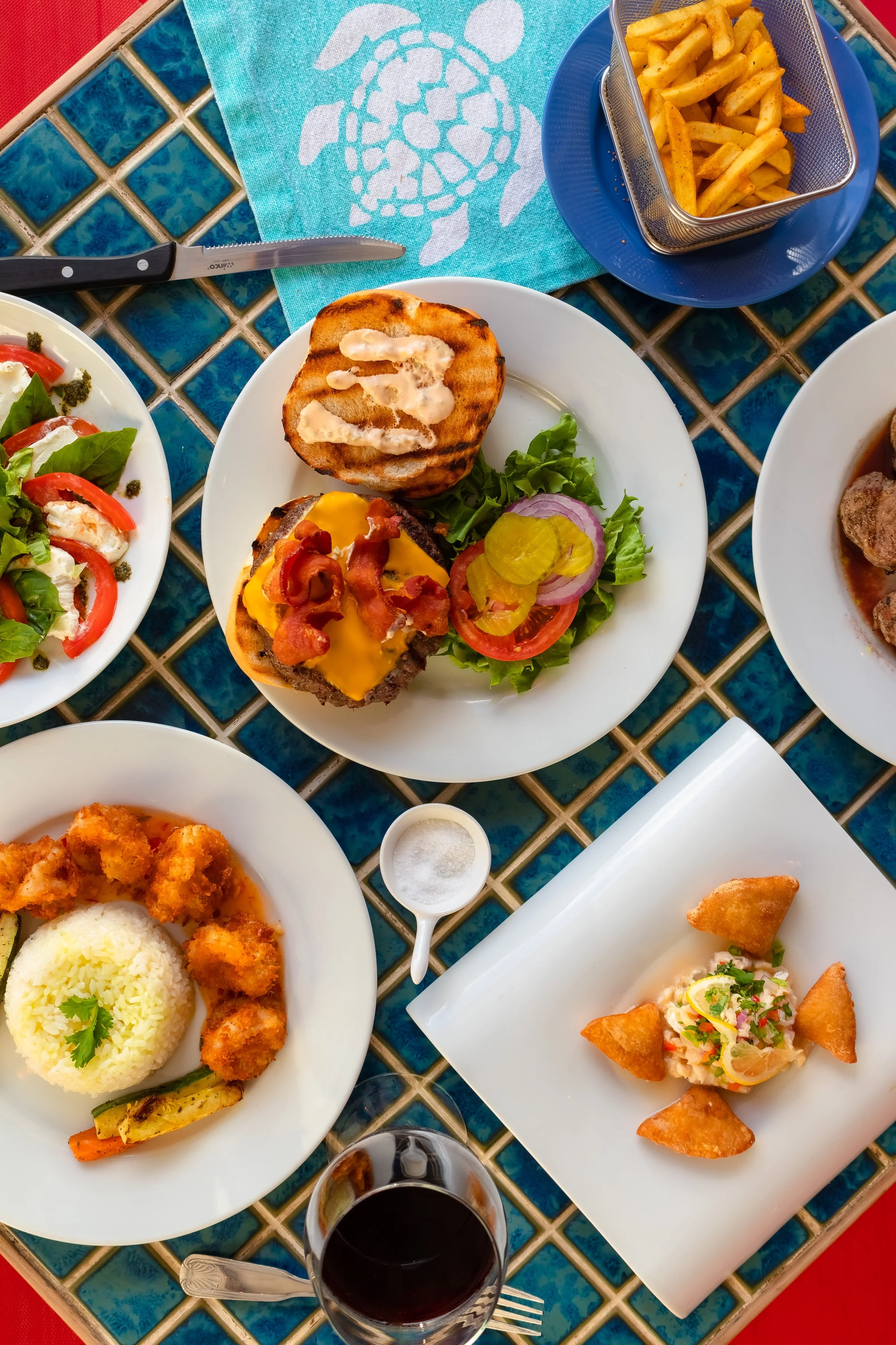 A colorful meal spread out on a table with dishes including fries, a burger, fish, rice, fried chicken, and a salad, along with a glass of red wine and a salt shaker.