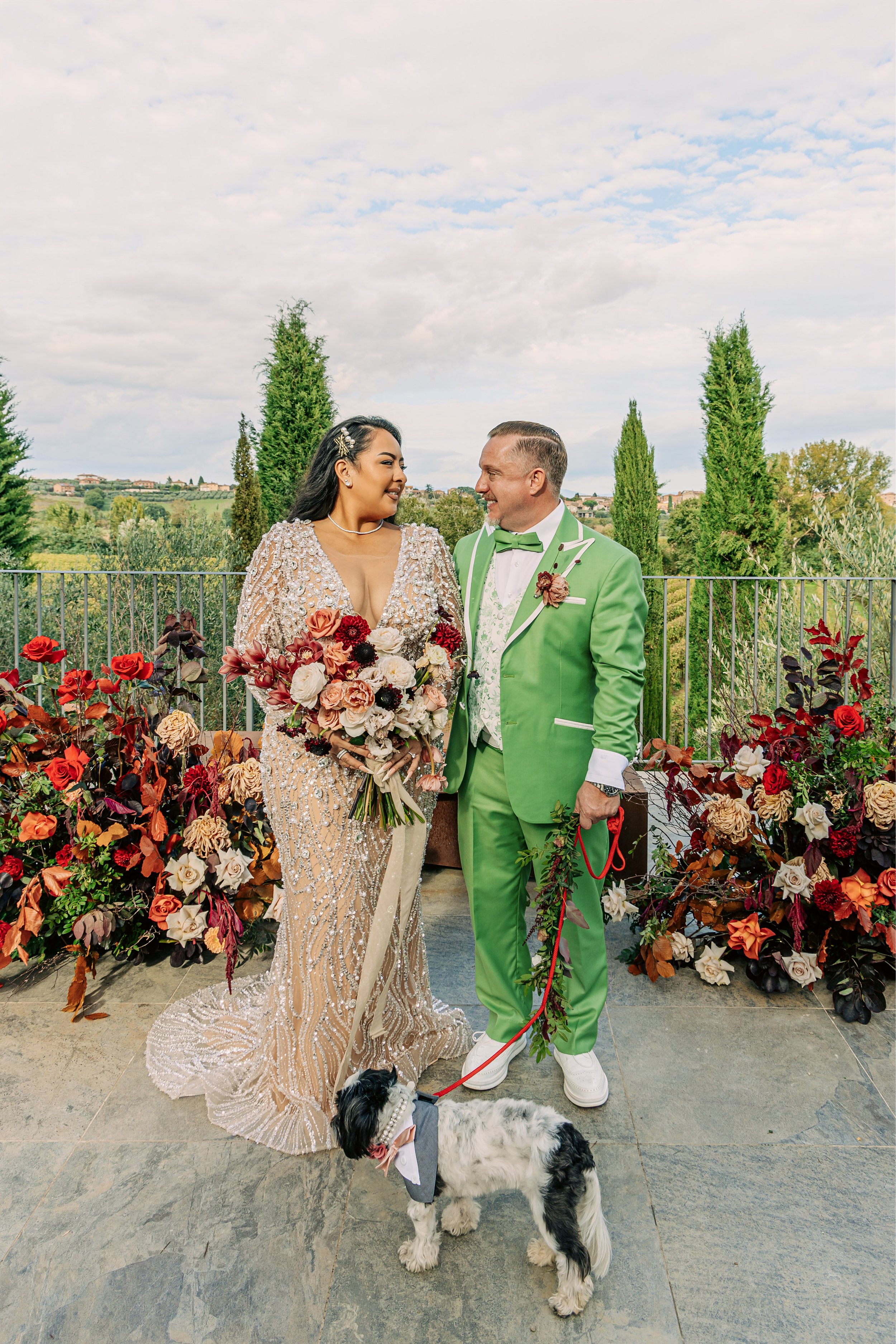 A bride and groom standing together surrounded by vibrant flowers and greenery outdoors. The bride is wearing an intricate, sheer, beaded gown, and holding a large bouquet of flowers. The groom is dressed in a bright green suit with a bow tie and bou
