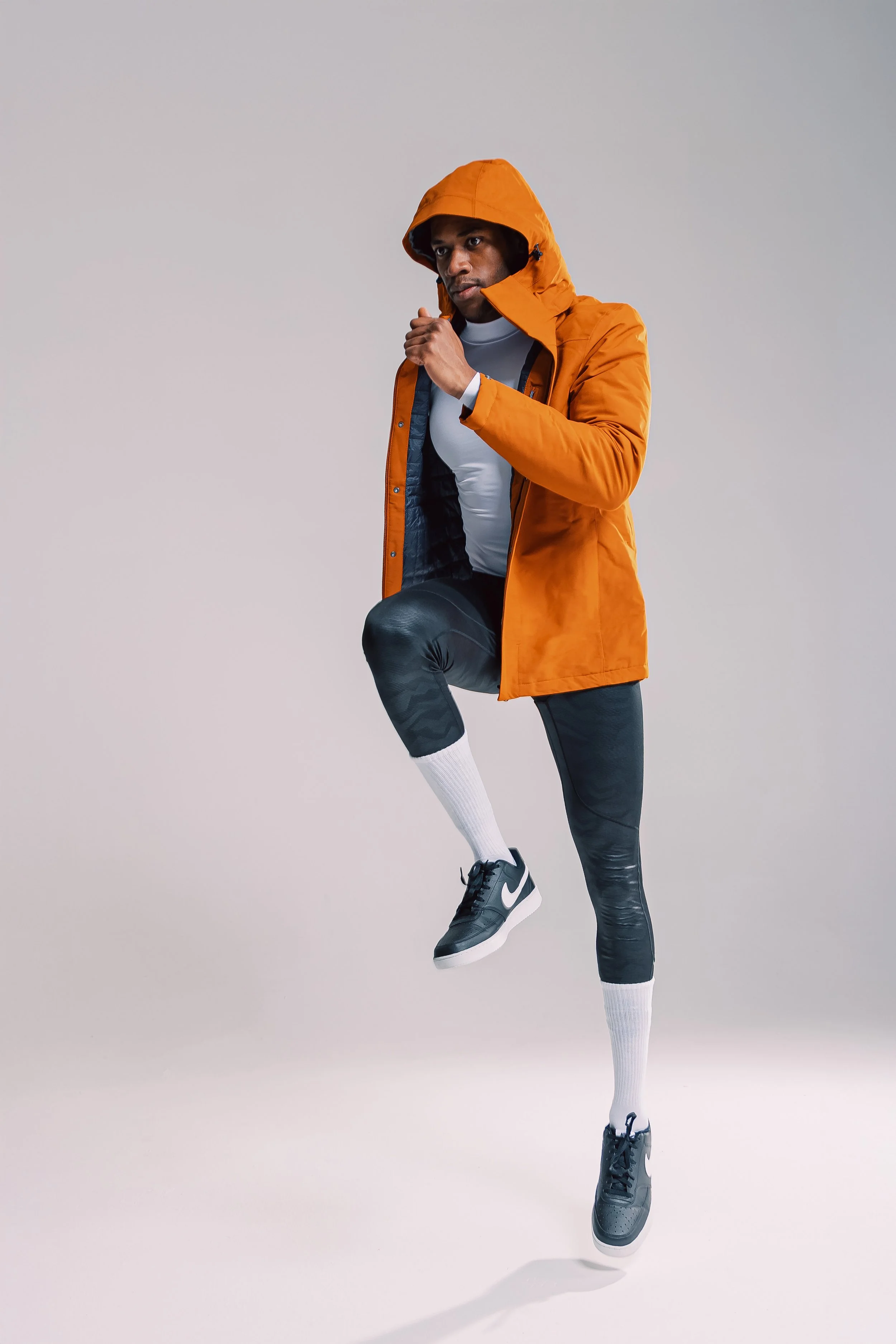 A man wearing a bright orange hooded jacket, black athletic leggings, and sneakers jumps with one knee bent and one arm raised against a plain gray background.