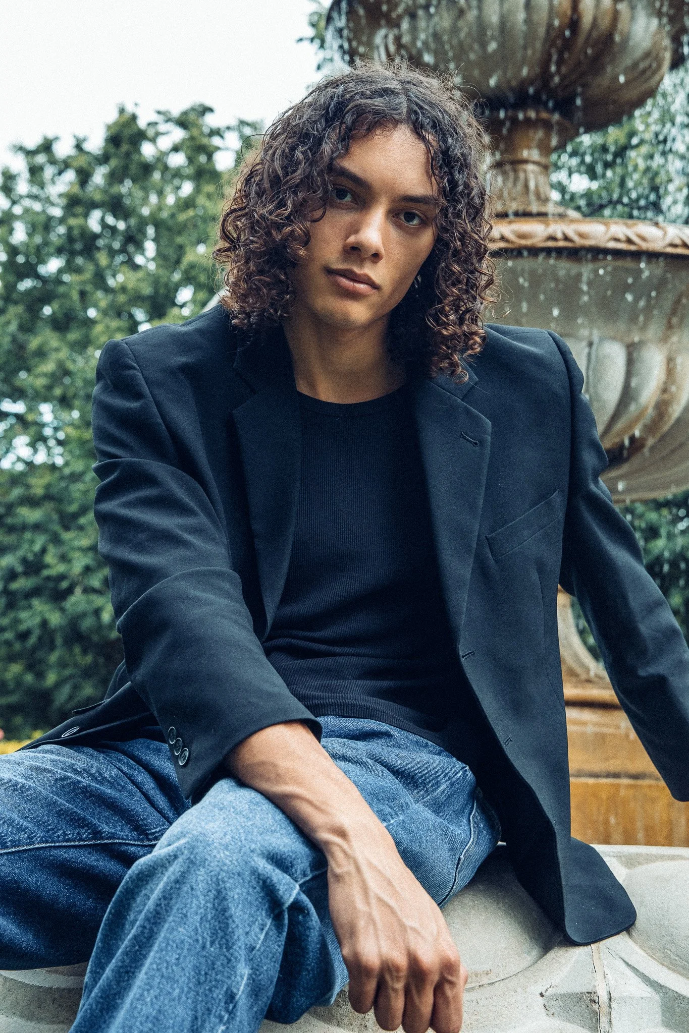 A young person with curly hair and light skin sitting outdoors in front of a fountain with trees in the background, wearing a dark blazer over a black shirt and blue jeans.
