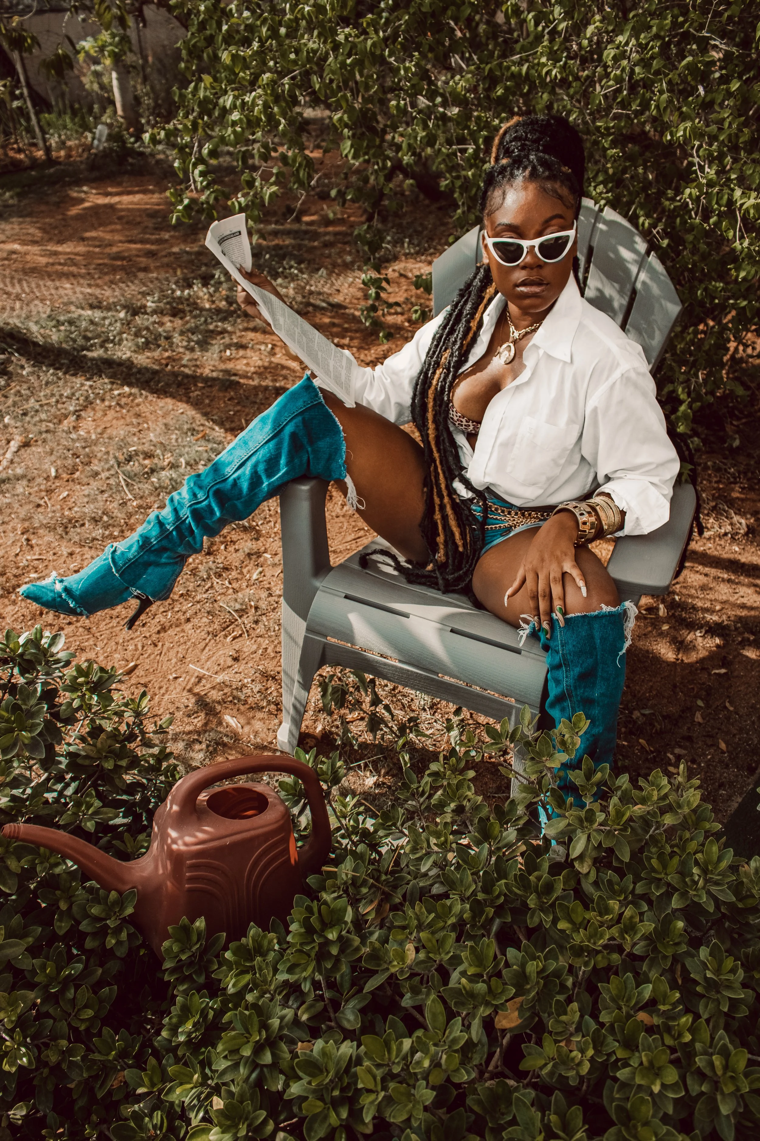 A woman with long braided hair wearing sunglasses, a white shirt, and thigh-high ripped jeans, sitting on a gray outdoor chair surrounded by greenery, reading a newspaper with a brown watering can nearby.