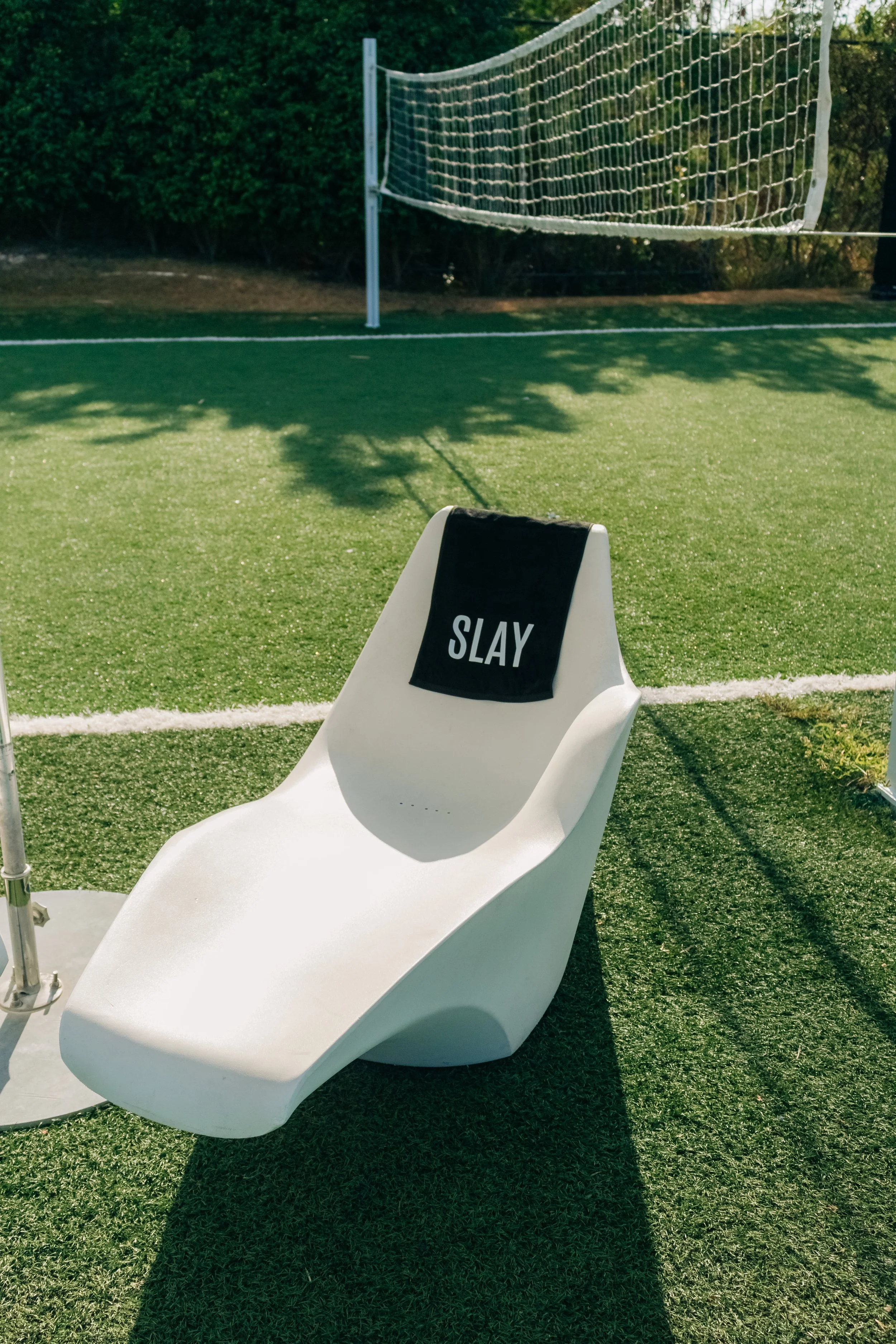 A white lounge chair with a black towel that says 'SLAY' on it, positioned on a green artificial turf field near a volleyball net.