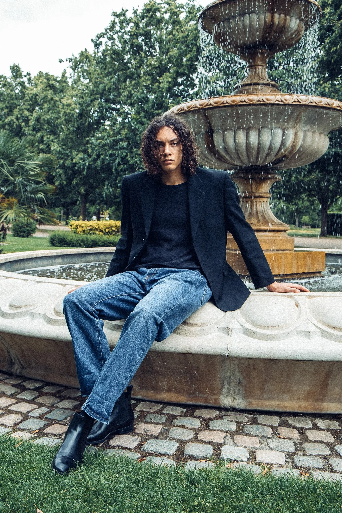 A person with shoulder-length curly hair sitting on the edge of a fountain in a park, wearing a black blazer, a black top, loose blue jeans, and black boots. The fountain has water cascading from the top and is surrounded by greenery and trees.