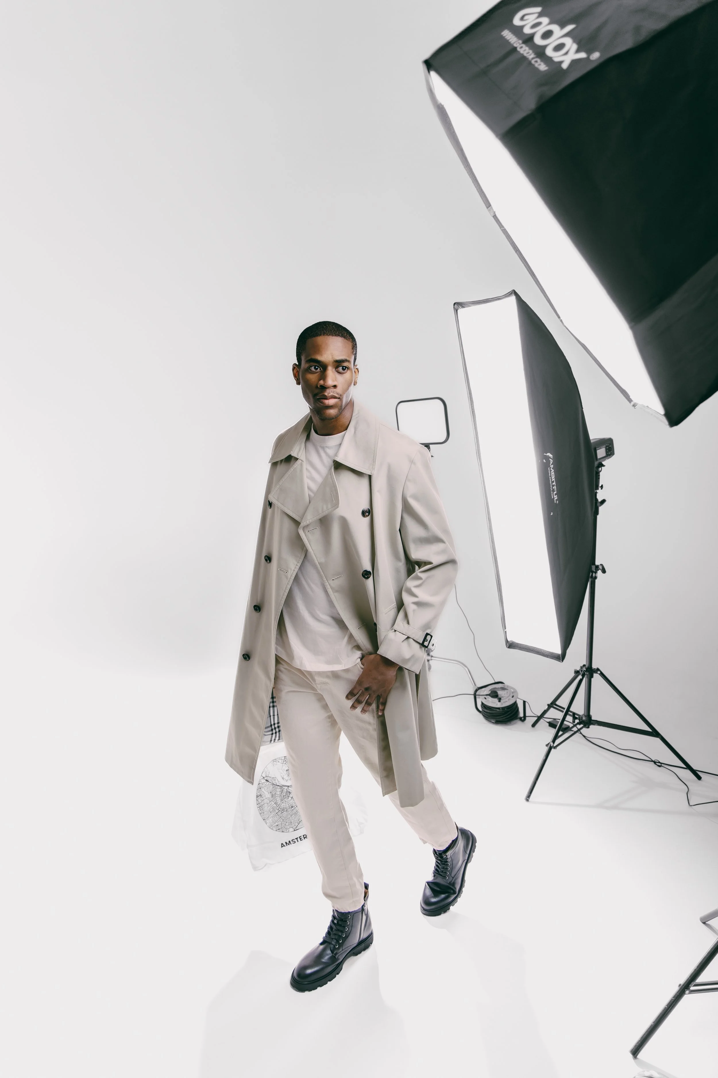 A man stands in a photography studio with professional lighting equipment, wearing a beige trench coat, white shirt, beige pants, and black boots.