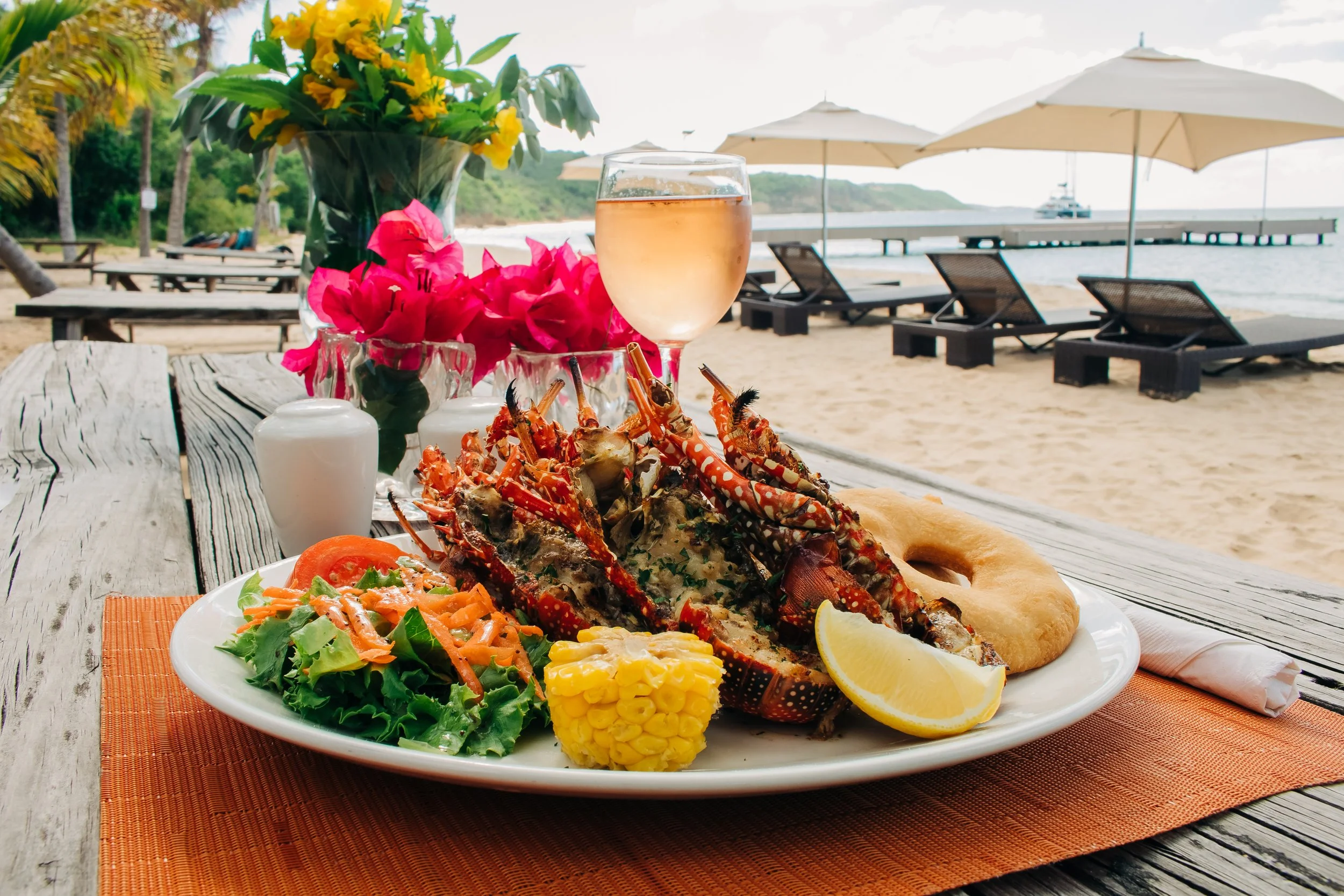 Plate of food with lobster tail, corn on the cob, salad, and bread, set on a wooden picnic table with orange placemat, at the beach.