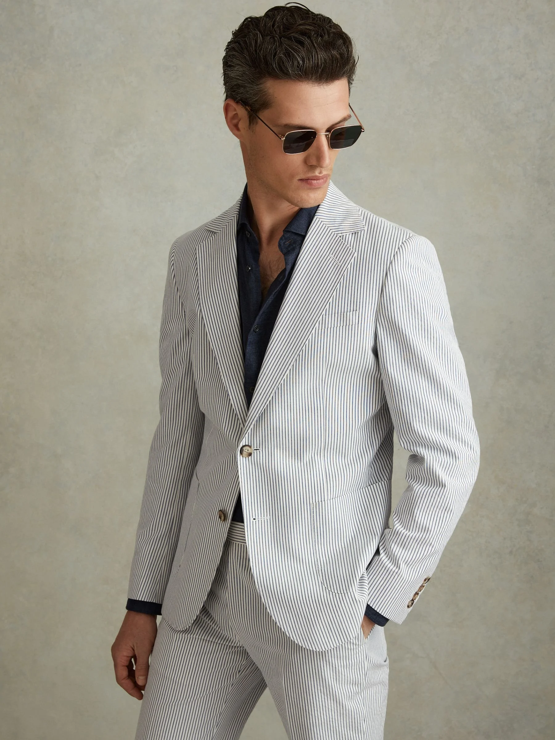 Man in a light-colored, pinstriped suit, black shirt, and sunglasses, standing against a neutral background.