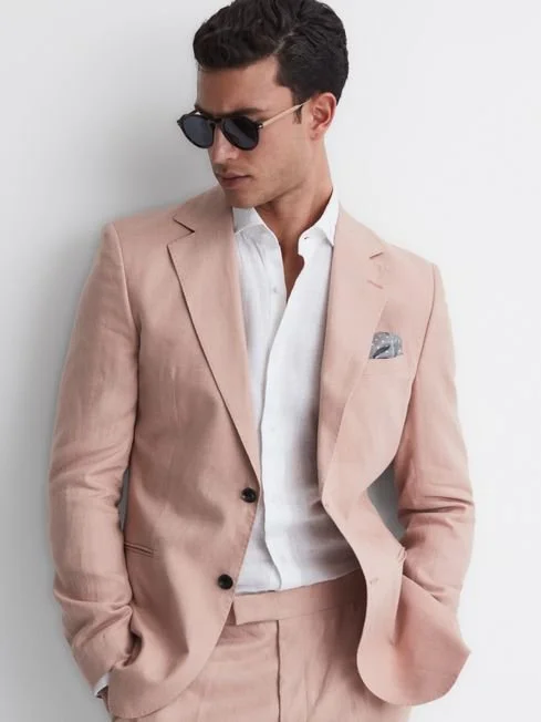 Man wearing a pink suit, white shirt, black sunglasses, and pocket square, standing against a white background.