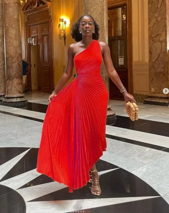 A woman in a red pleated one-shoulder evening gown standing in an ornate, elegant lobby with marble floors and gold-colored walls, holding a beaded clutch and wearing high-heeled sandals.