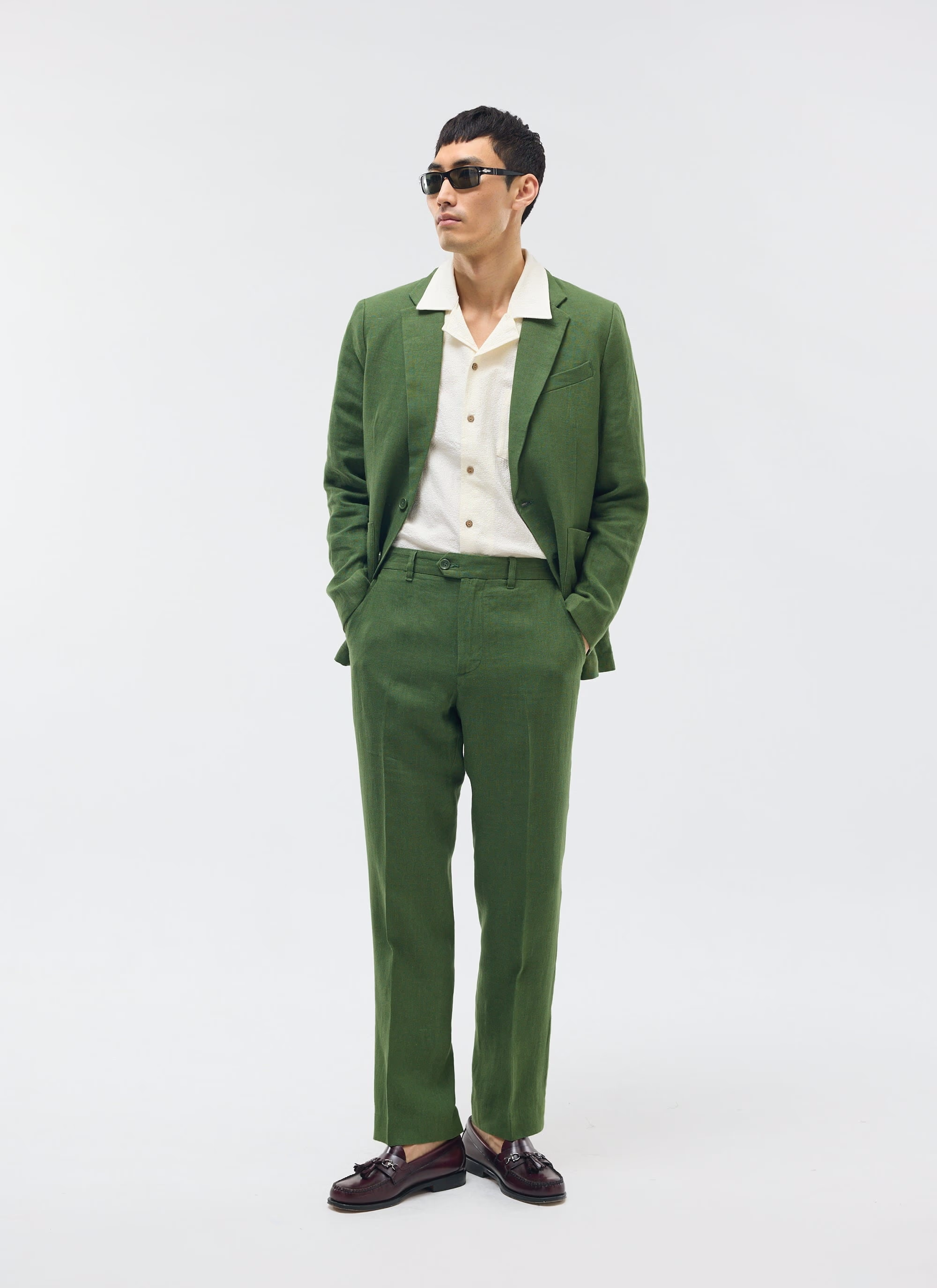 A young man wearing a green suit with a beige shirt, black sunglasses, and dark brown shoes, standing with hands in pockets against a plain white background.