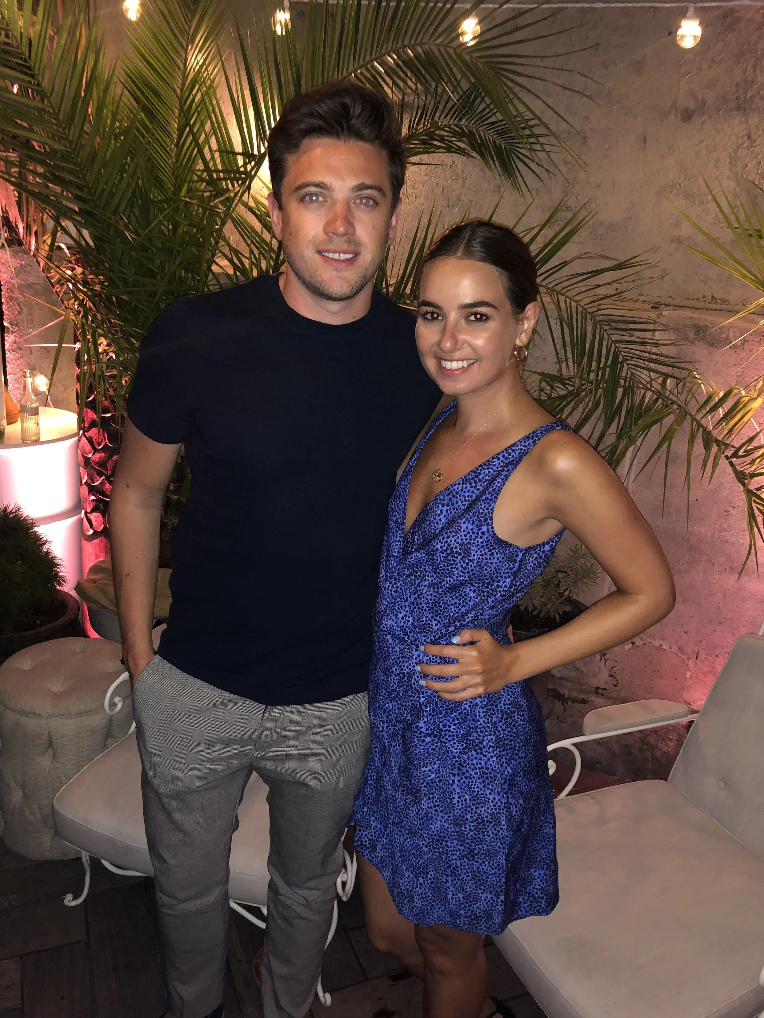 A man and a woman standing close together, smiling at the camera, in a tropical-themed setting with green plants and hanging string lights, with chairs and foliage in the background.