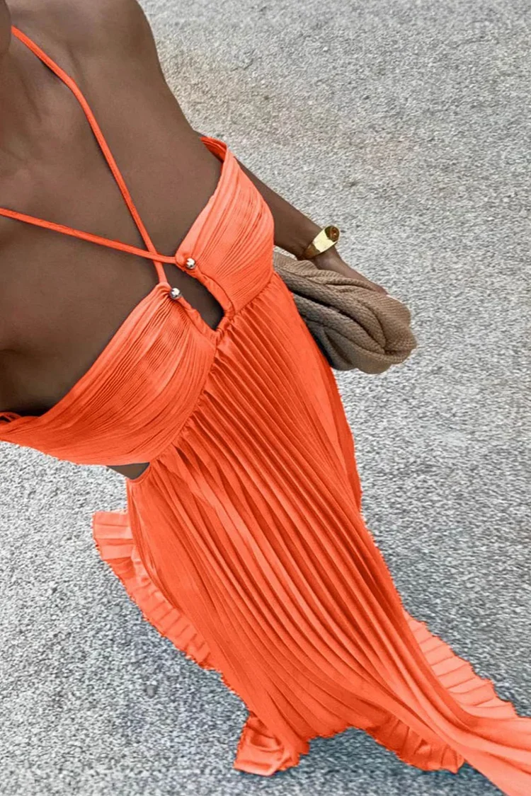 A woman wearing an orange pleated dress taking a selfie with a gray carpeted floor in the background, holding a beige clutch purse, and wearing a gold watch.
