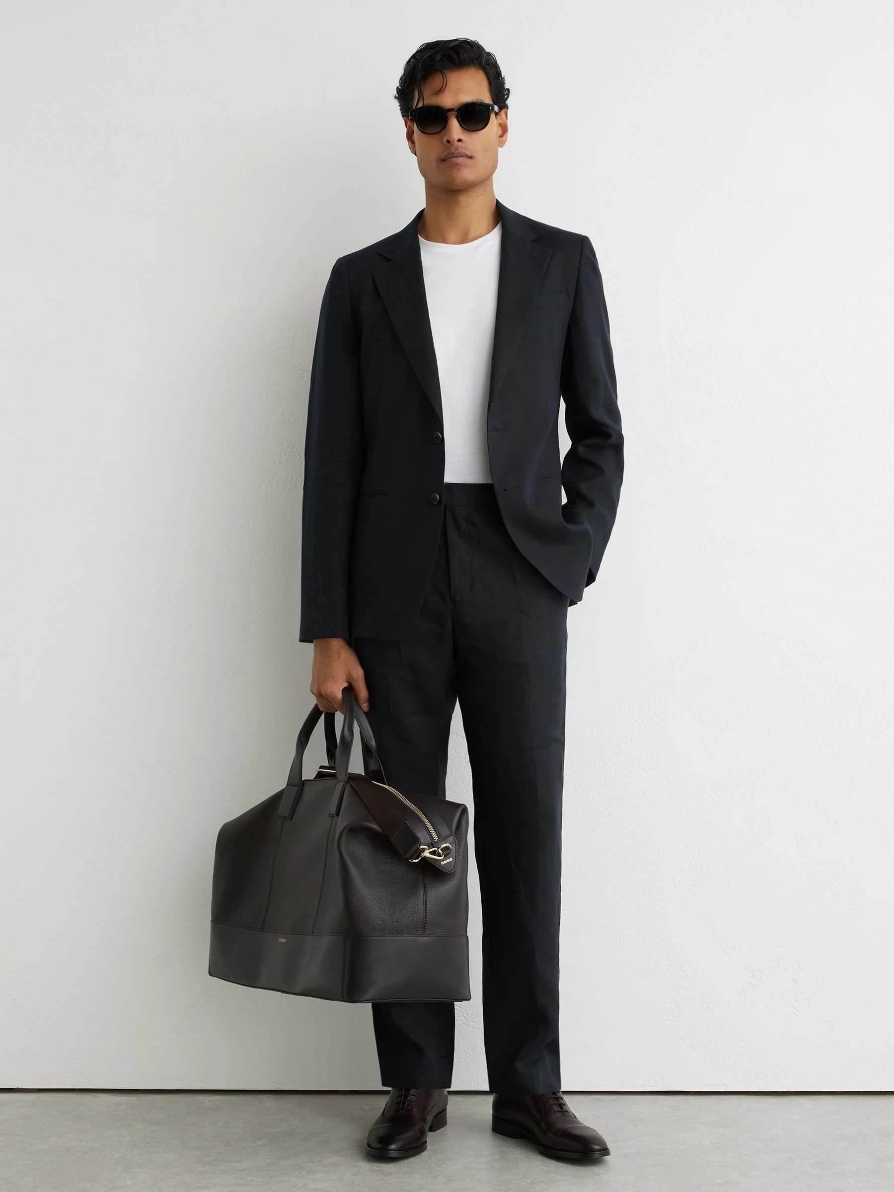 A man dressed in a black suit with a white shirt, wearing sunglasses, holding a large black leather duffel bag, standing against a plain white wall.