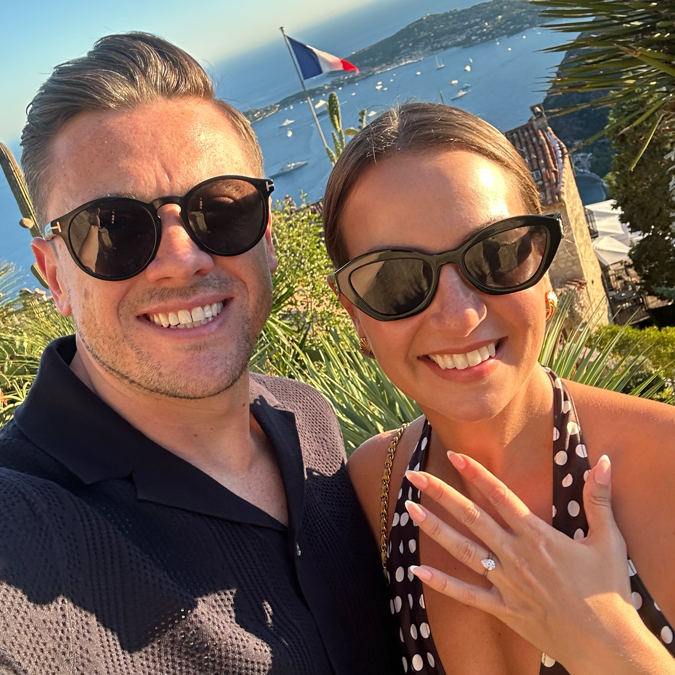 A smiling couple wearing sunglasses taking a selfie outdoors near a body of water, with boats, a French flag, and lush greenery in the background.