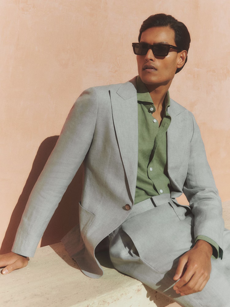 Man wearing sunglasses, green shirt, and gray suit, sitting against a peach-colored wall, casting a shadow.