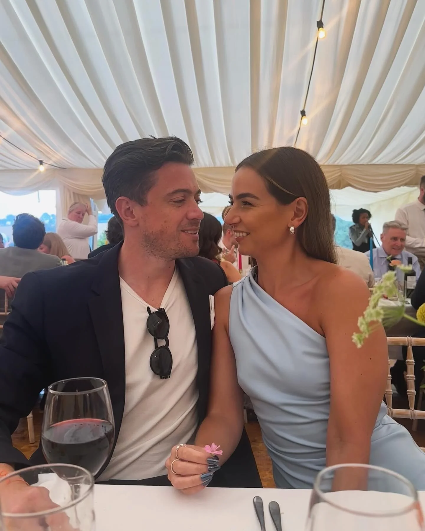 A man and woman smiling at each other at a wedding reception or formal event inside a decorated tent, with tables, wine glasses, and other guests in the background.