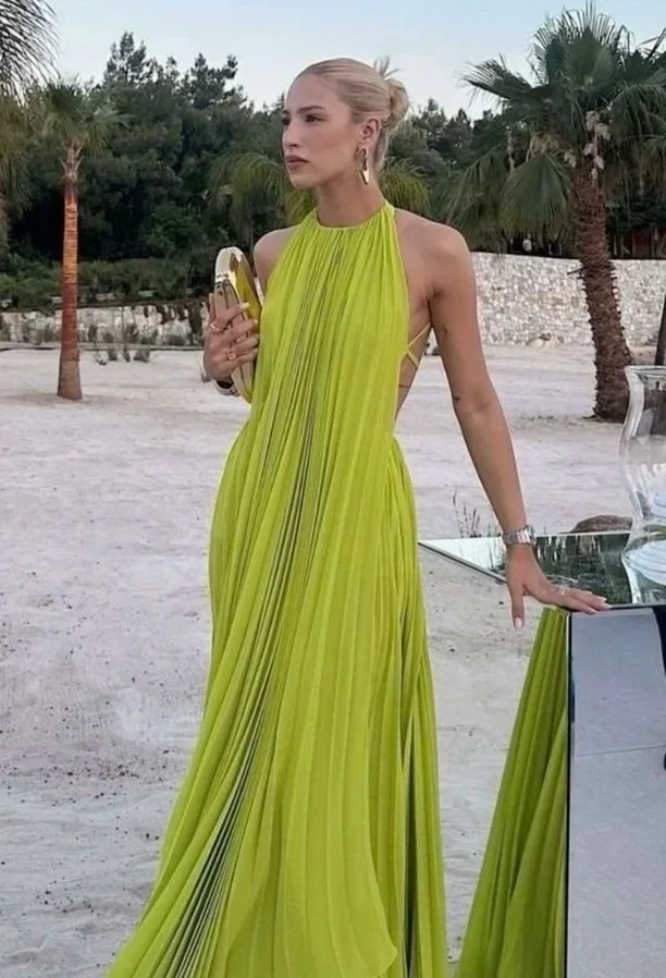 woman in a bright yellow pleated gown standing outdoors on sandy ground with palm trees in the background, holding a banana and a phone.