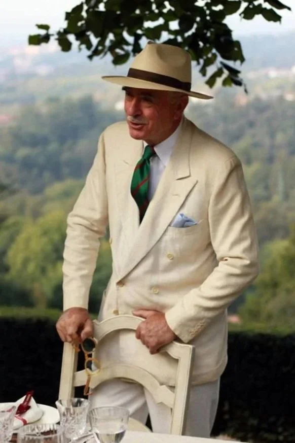 A man dressed in a cream-colored suit and hat, with a green and red striped tie, standing at a table outdoors with a scenic landscape in the background.