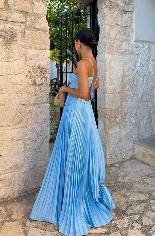 A woman in a long, pleated blue dress standing outside near a stone wall and black gate, holding a beige clutch purse.