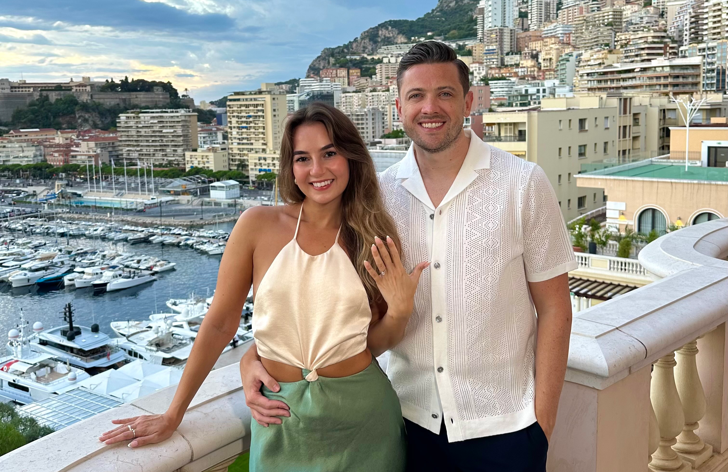 A smiling couple standing on a balcony overlooking a marina with yachts, with a cityscape and hillside in the background.
