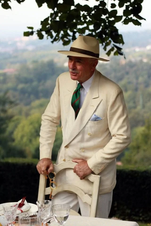 An elderly man dressed in a cream-colored suit with a white shirt, a green and red tie, and a pocket square, holding a chair behind a table set with glasses, outdoors with a landscape of greenery in the background, and wearing a beige fedora hat with a black band.