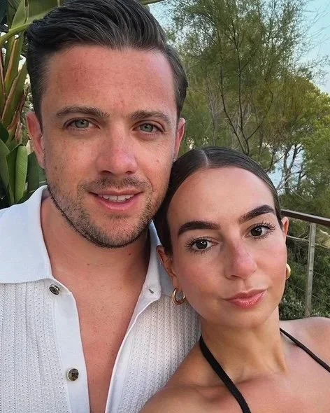 A man and a woman taking a selfie outdoors, with trees and a railing in the background.