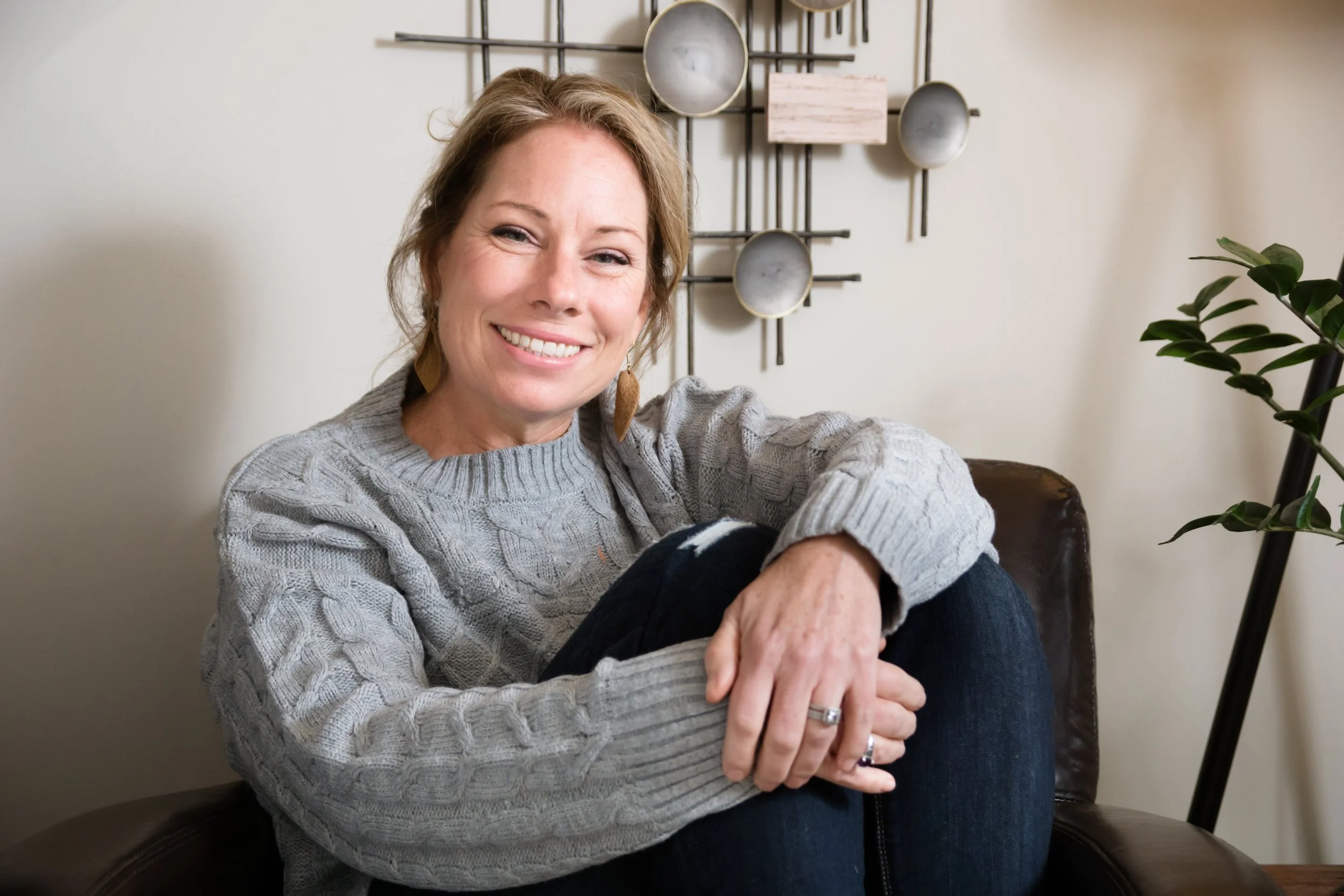A woman with blonde hair in a gray sweater sitting on a chair, smiling at the camera, with a decorative wall hanging and a plant in the background.