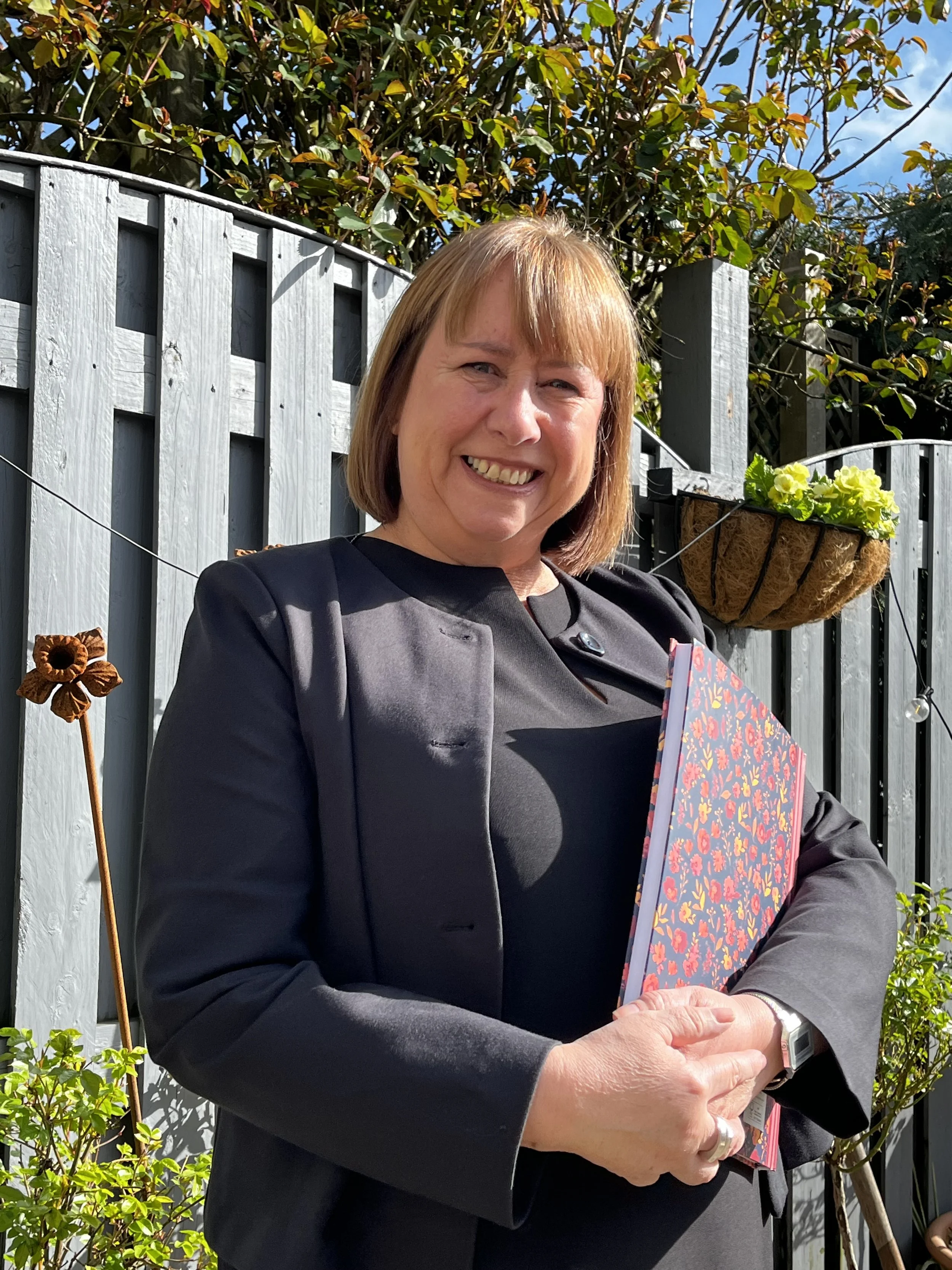 A smiling woman with short brown hair and a black outfit holding a floral patterned folder, standing outdoors in front of a gray wooden fence, with green plants and a hanging basket with green flowers behind her on a sunny day.