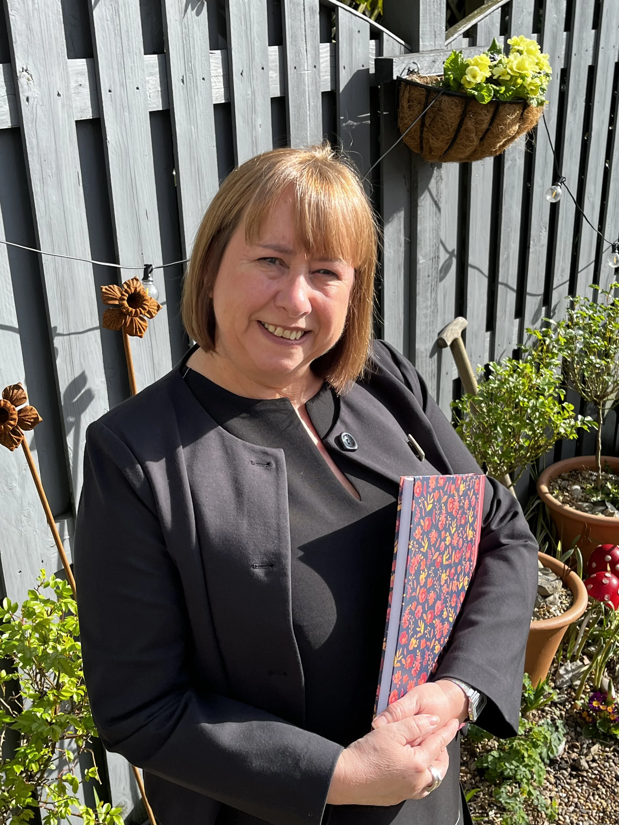 Woman with shoulder-length light brown hair smiling outdoors, holding a colorful floral notebook, standing in a garden with potted plants, a gray wooden fence, and decorative flower stakes.